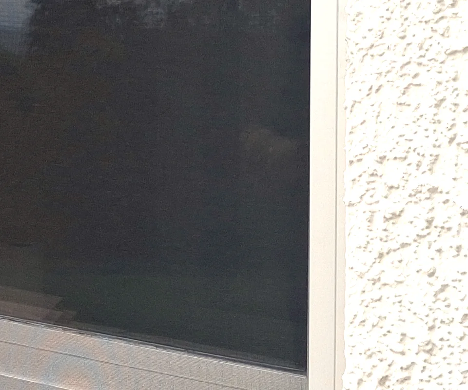 Moustiquaire latérale enroulable tirée devant une fenêtre, toile sombre tendue dans ses profils, mur crépi clair.