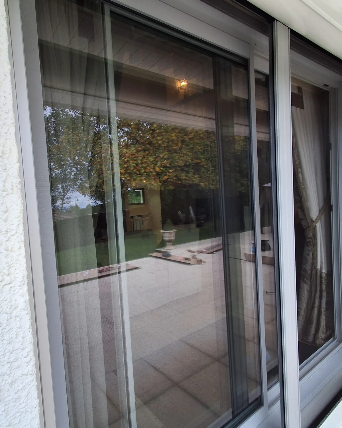 Baie coulissante en aluminium blanc d’une maison de Vétraz-Monthoux, équipée d’une moustiquaire latérale enroulable, dans une façade au crépi blanc ; la terrasse et le jardin se reflètent dans la vitre.
