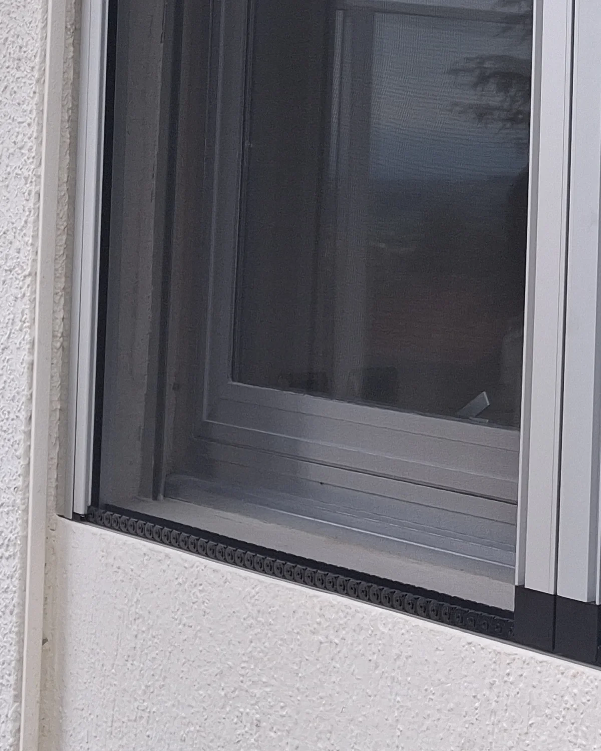 Bas d’une fenêtre de Vétraz-Monthoux équipée d’une moustiquaire latérale enroulable : le profil vertical de la cassette et le rail au sol à brosse noire, sur une façade au crépi blanc.