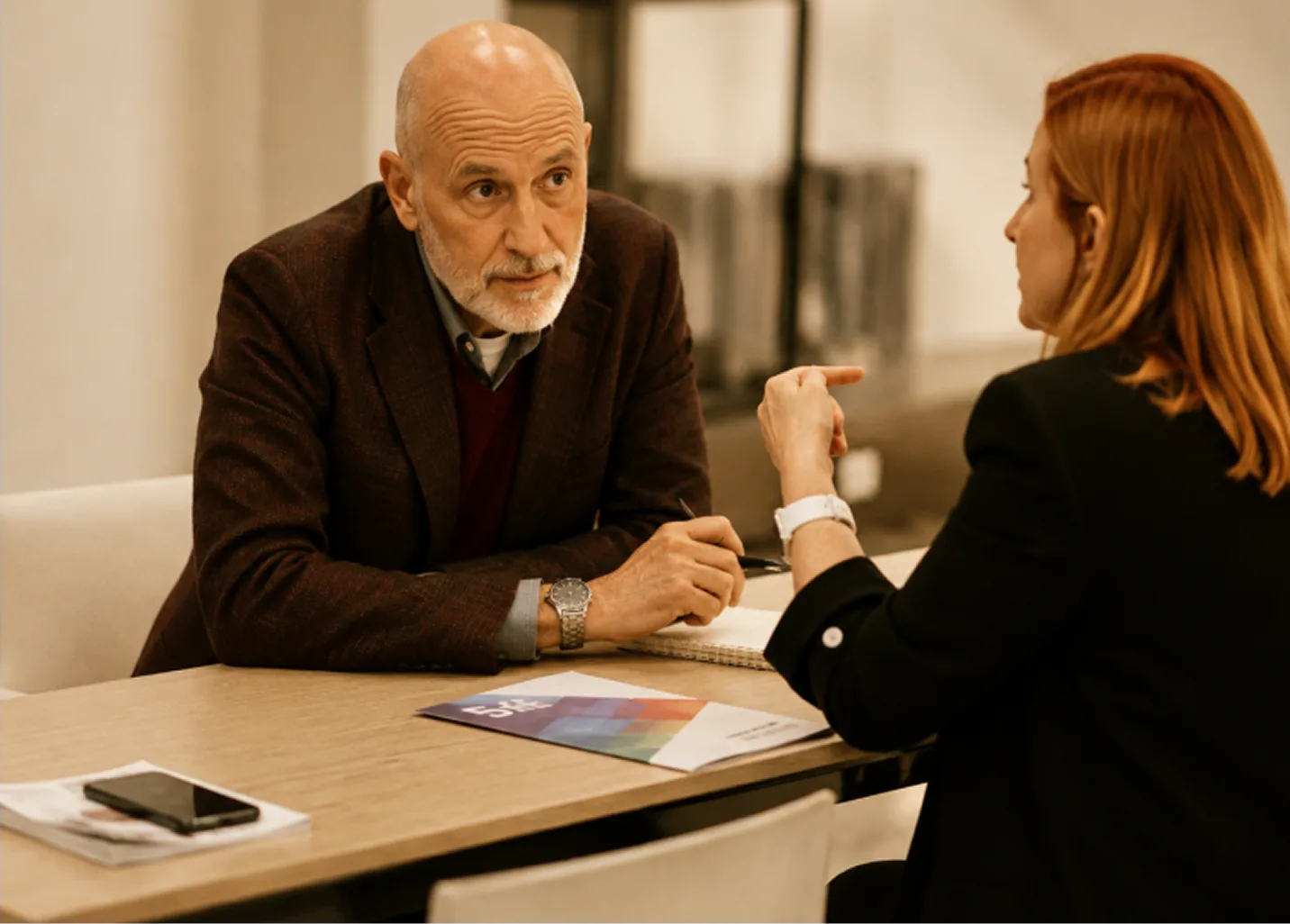 An older man in a burgundy jacket talking with a woman across a meeting table