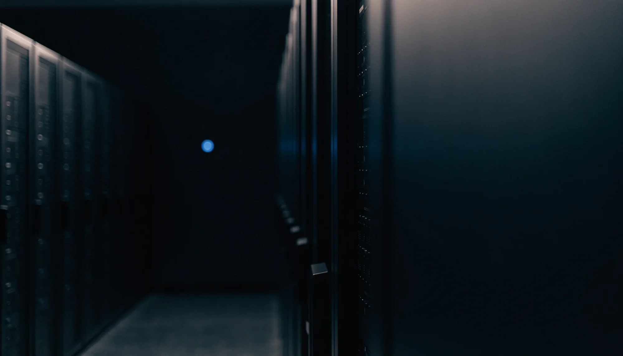 Dunkler Serverraum mit Reihen schwarzer Server-Racks und einem einzelnen blauen Statuslicht in der Ferne