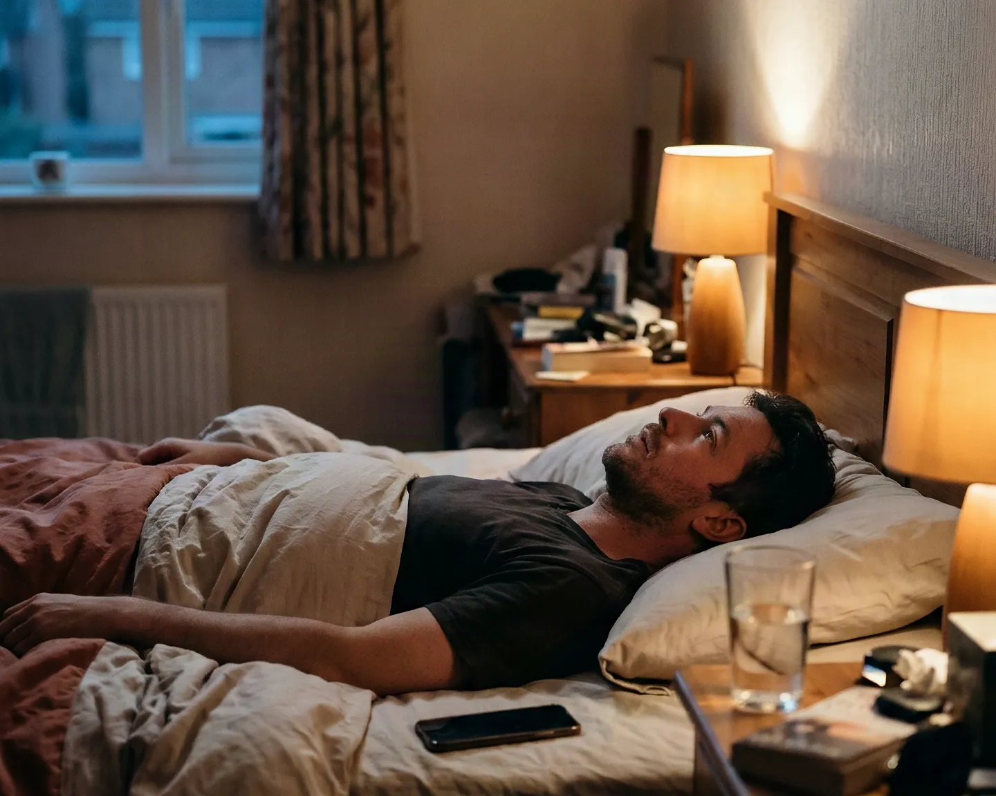 A man lying awake in bed at dusk, staring at the ceiling