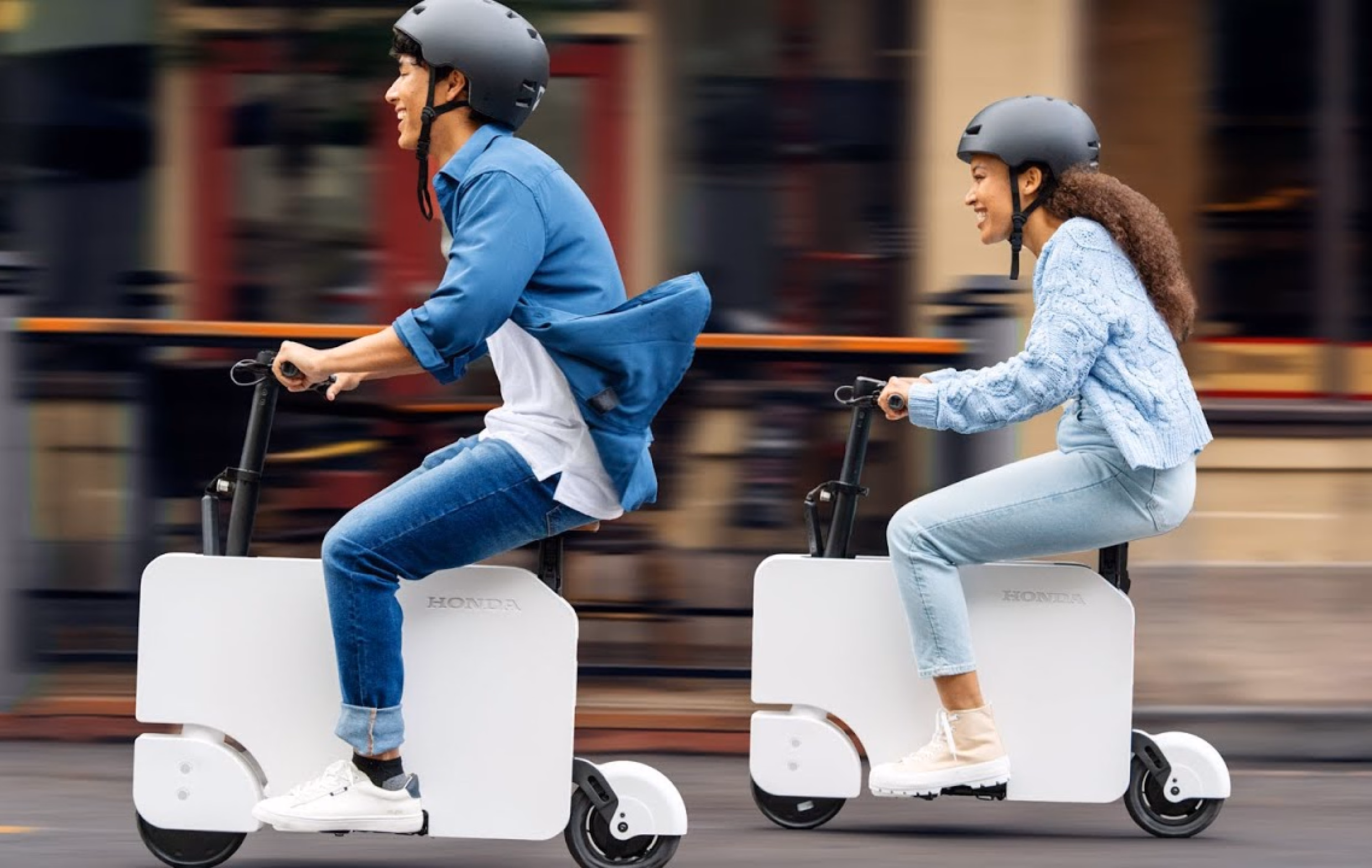 Two riders in helmets on white Honda Motocompacto folding electric scooters, moving along a city street