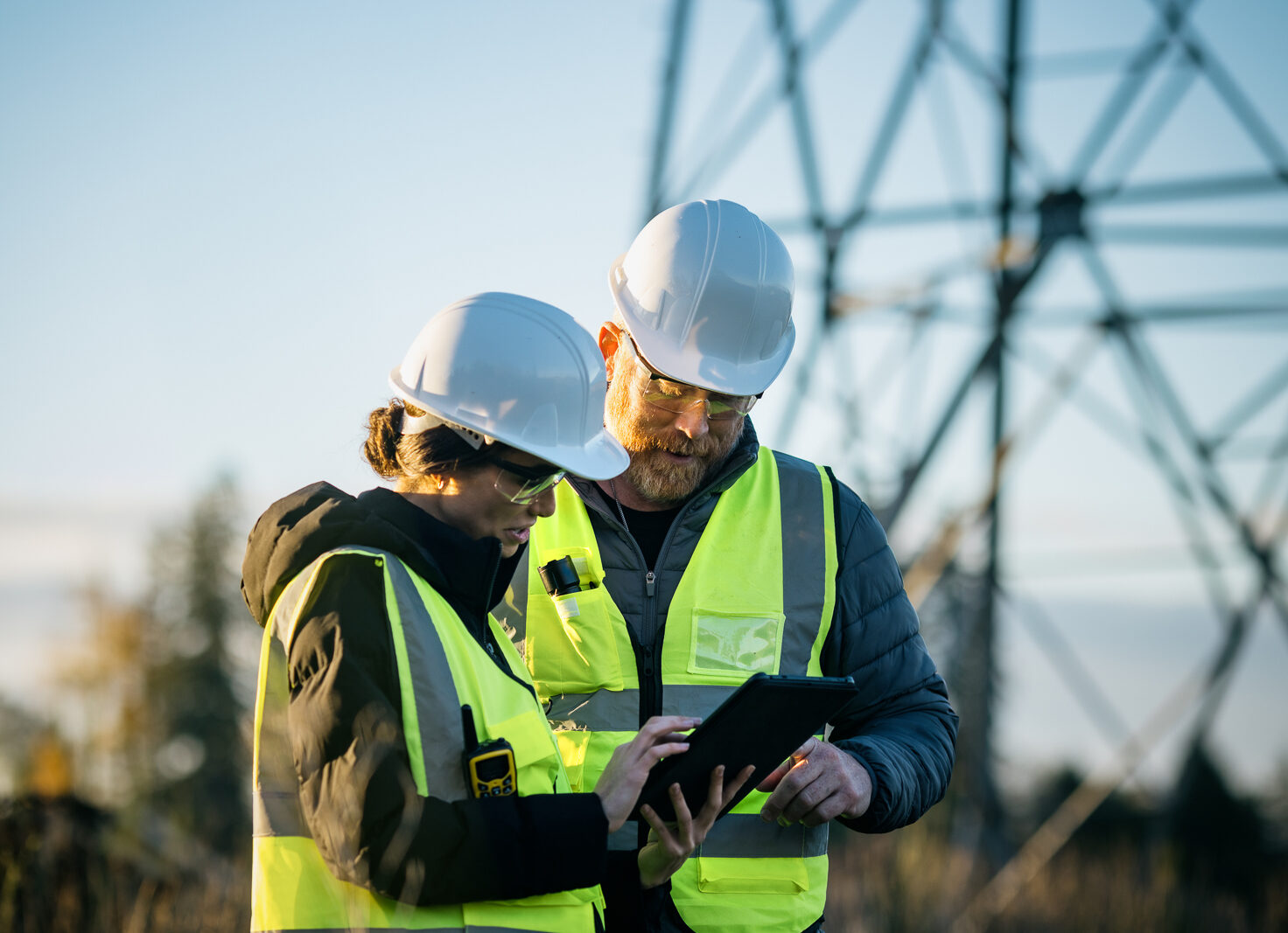 Two utility workers in hard hats and high-visibility vests reviewing a tablet beneath a power transmission tower