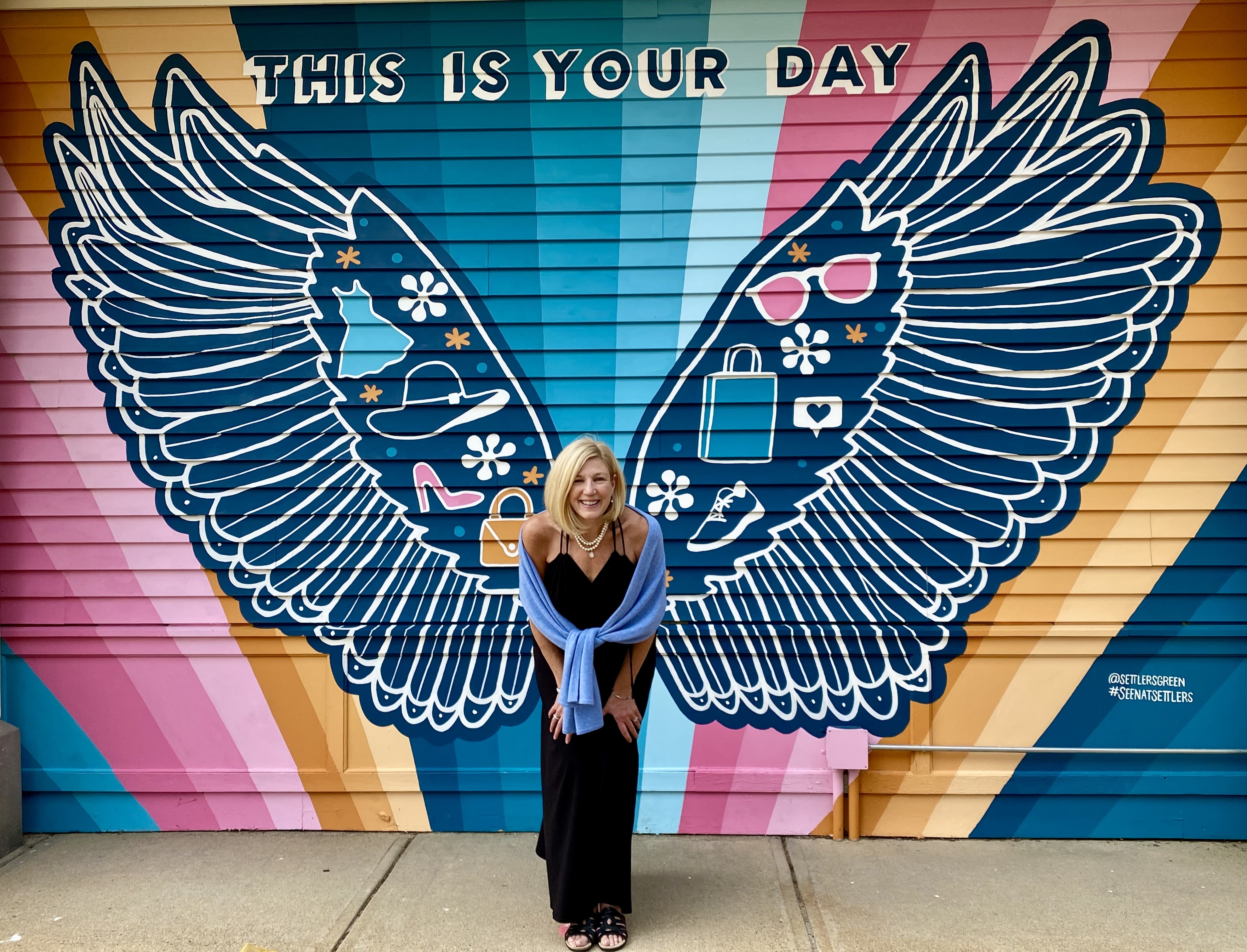 Kat Frati smiling in front of a colorful mural of wings that reads This Is Your Day