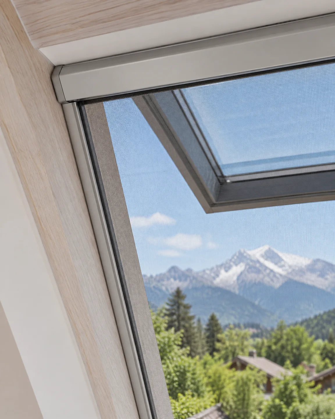 Vue rapprochée d’une moustiquaire de fenêtre de toit : cassette en aluminium clair en haut de l’embrasure en bois, coulisse le long de la joue, toile fine devant la fenêtre ouverte et les sommets enneigés.