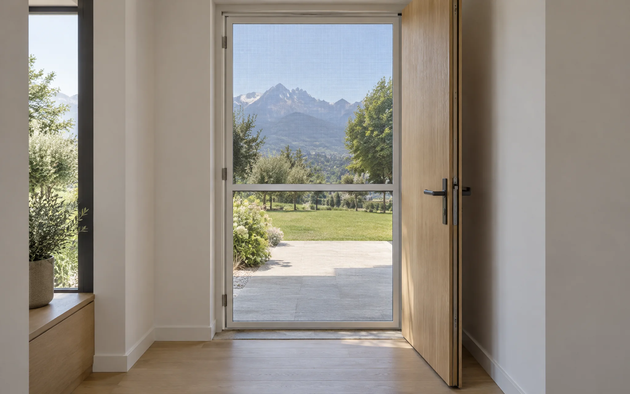 Entrée claire au parquet de chêne, porte d’entrée en bois ouverte et moustiquaire battante à cadre blanc fermée dans l’embrasure, jardin et montagnes derrière.