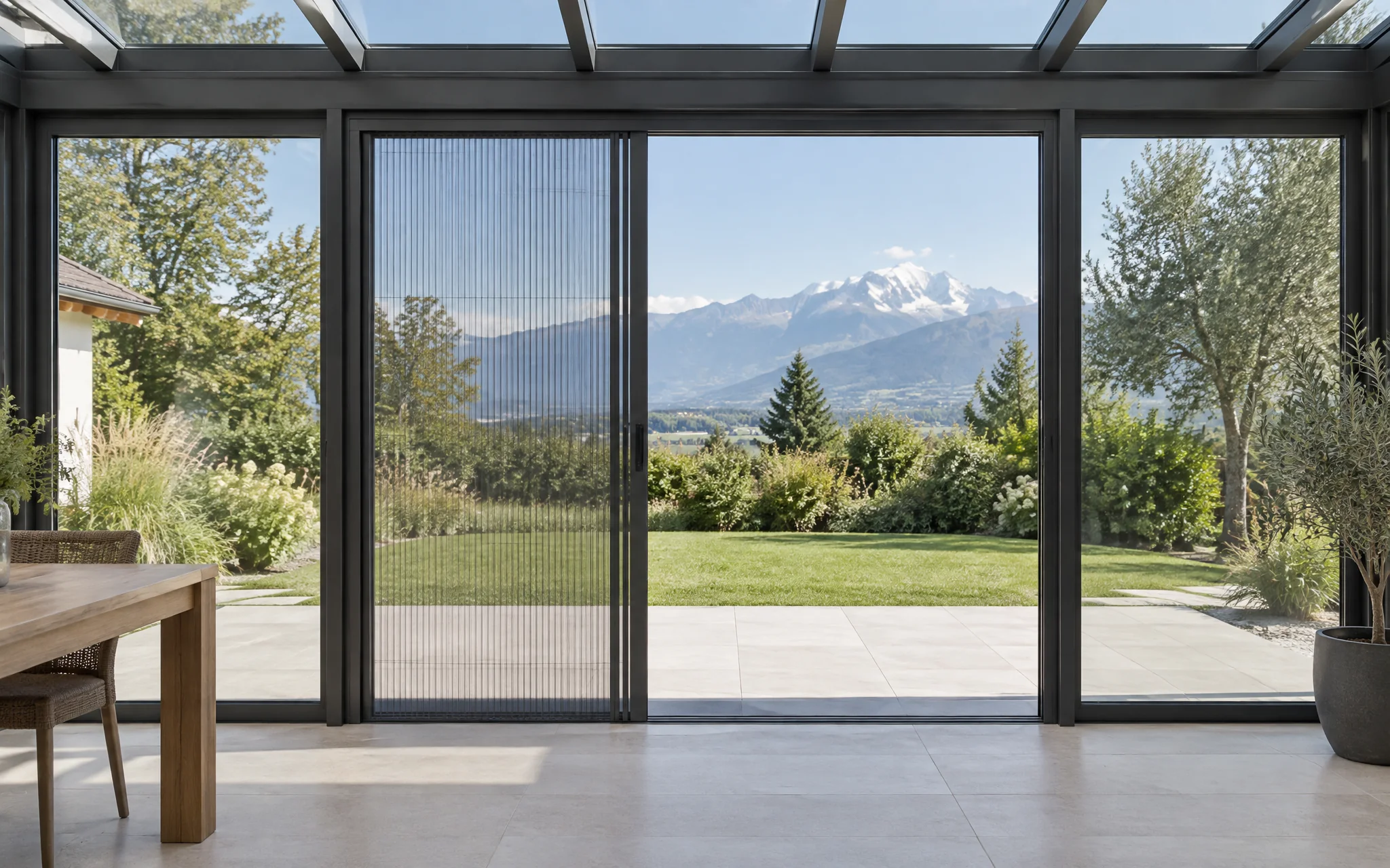 Véranda en aluminium anthracite ouverte sur le jardin, moustiquaire plissée tirée sur une partie du grand coulissant, Mont-Blanc au loin.