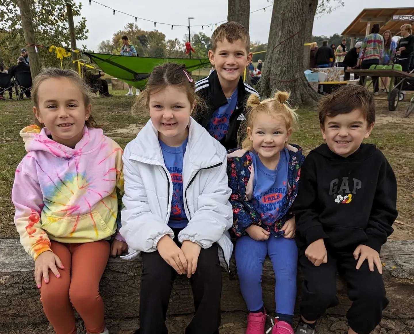 Four young kids sitting together on a log, two in tie-dye, smiling for the camera