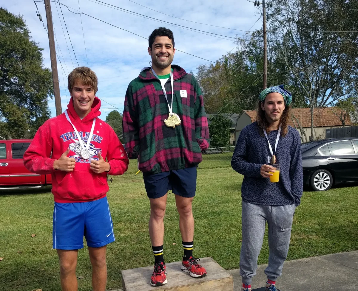 Three runners wearing medals on a grassy lawn, one standing on a wooden box like a podium