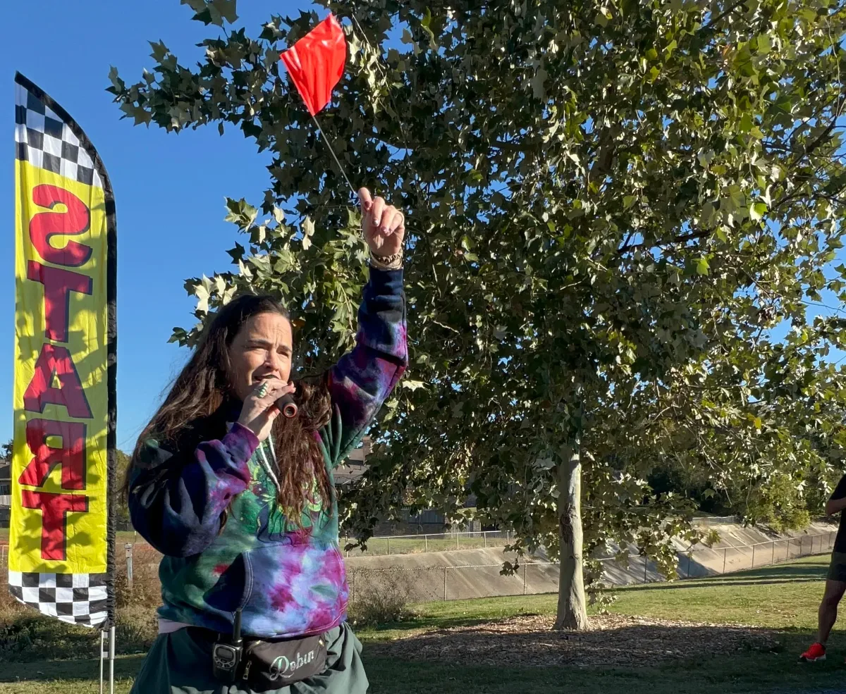 A woman in a tie-dye shirt raising a red flag beside the start banner to begin the trot