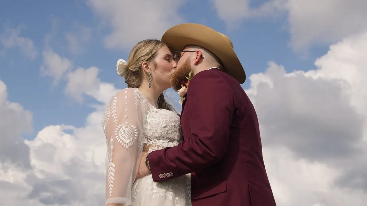 Kelly and Grant kissing beneath a bright, cloudy sky on their wedding day