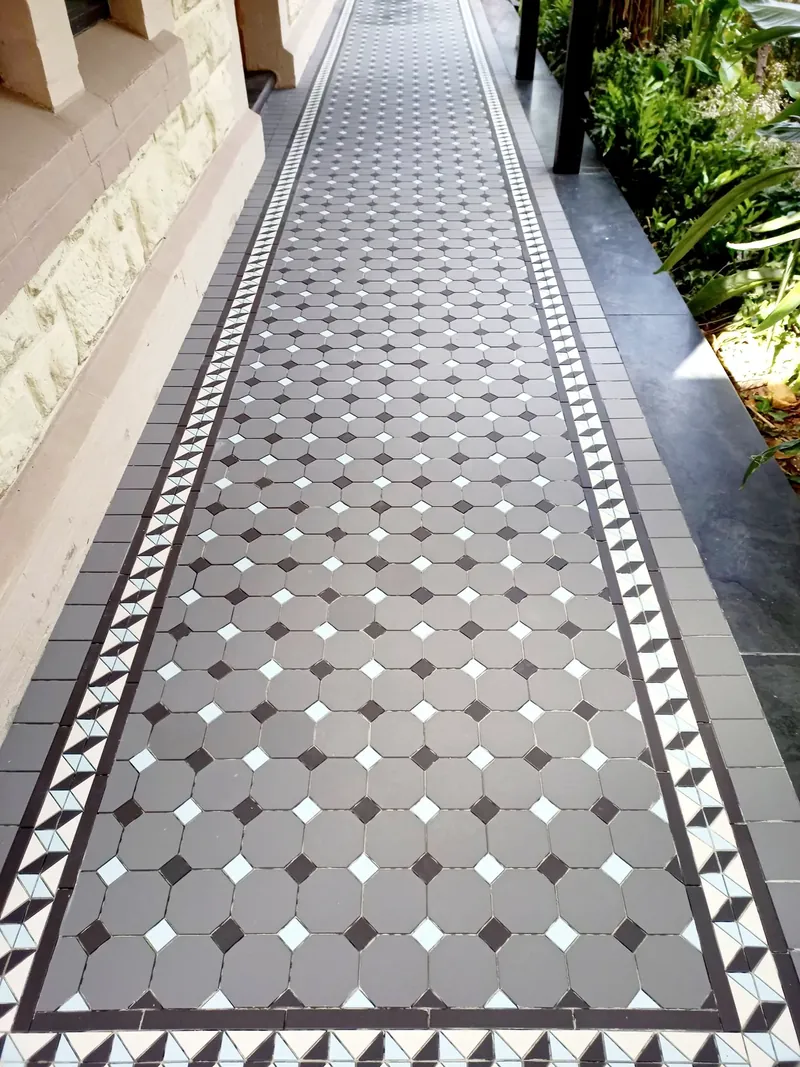 Path of geometric tessellated floor tiles with a decorative border beside a stone wall and garden