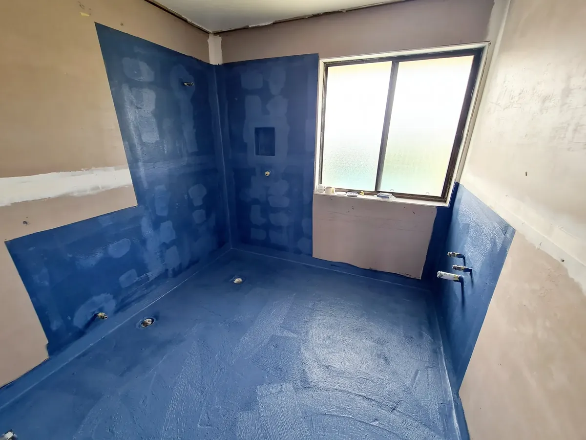 Bathroom walls and floor covered in blue waterproofing membrane with a window and a floor waste, before tiling