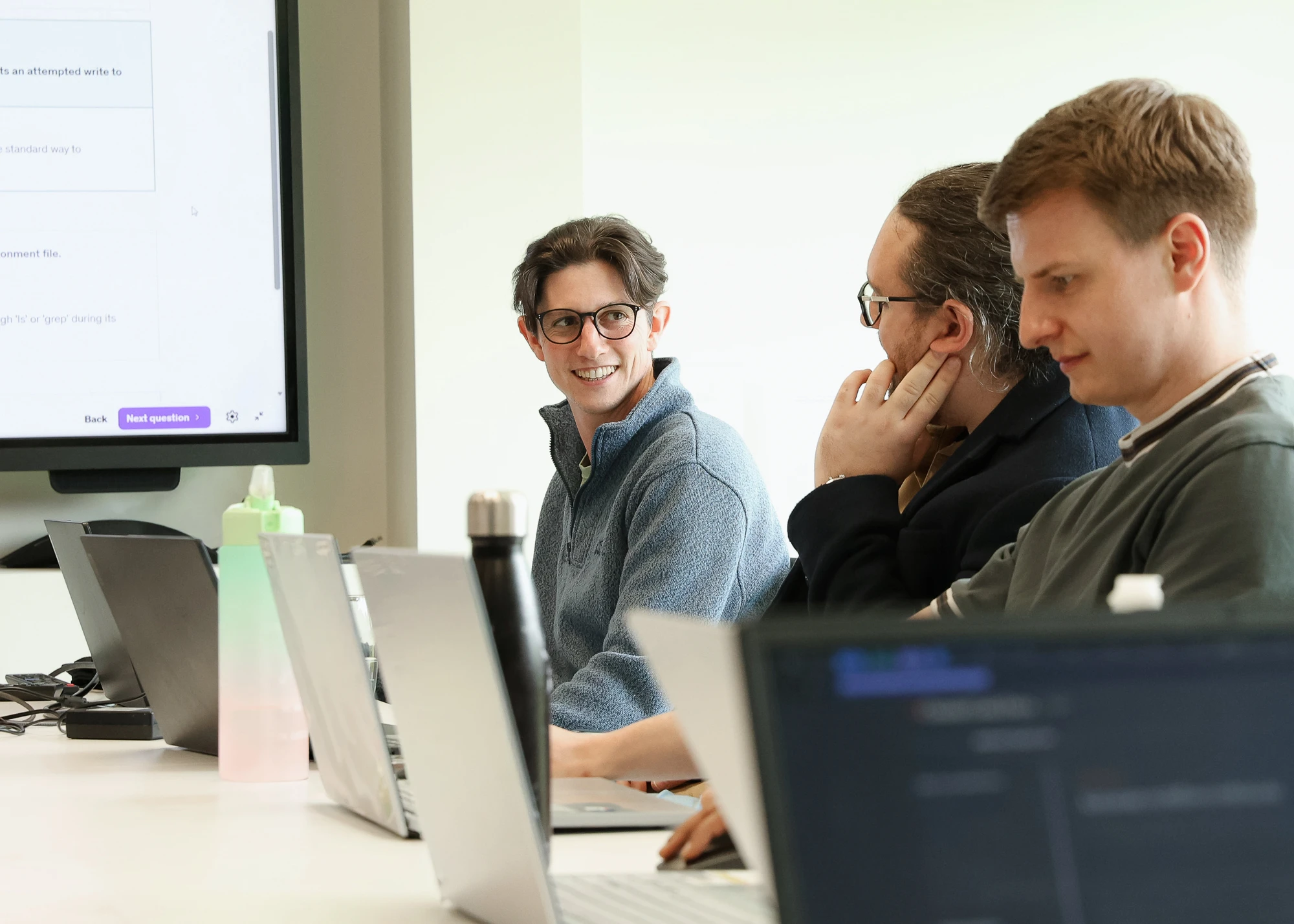 Three people sitting at a table with laptops, a large monitor on the wall displaying a user interface, and water bottles on the table; one person in the middle wearing glasses and a blue fleece is smiling and looking at the person on the right.