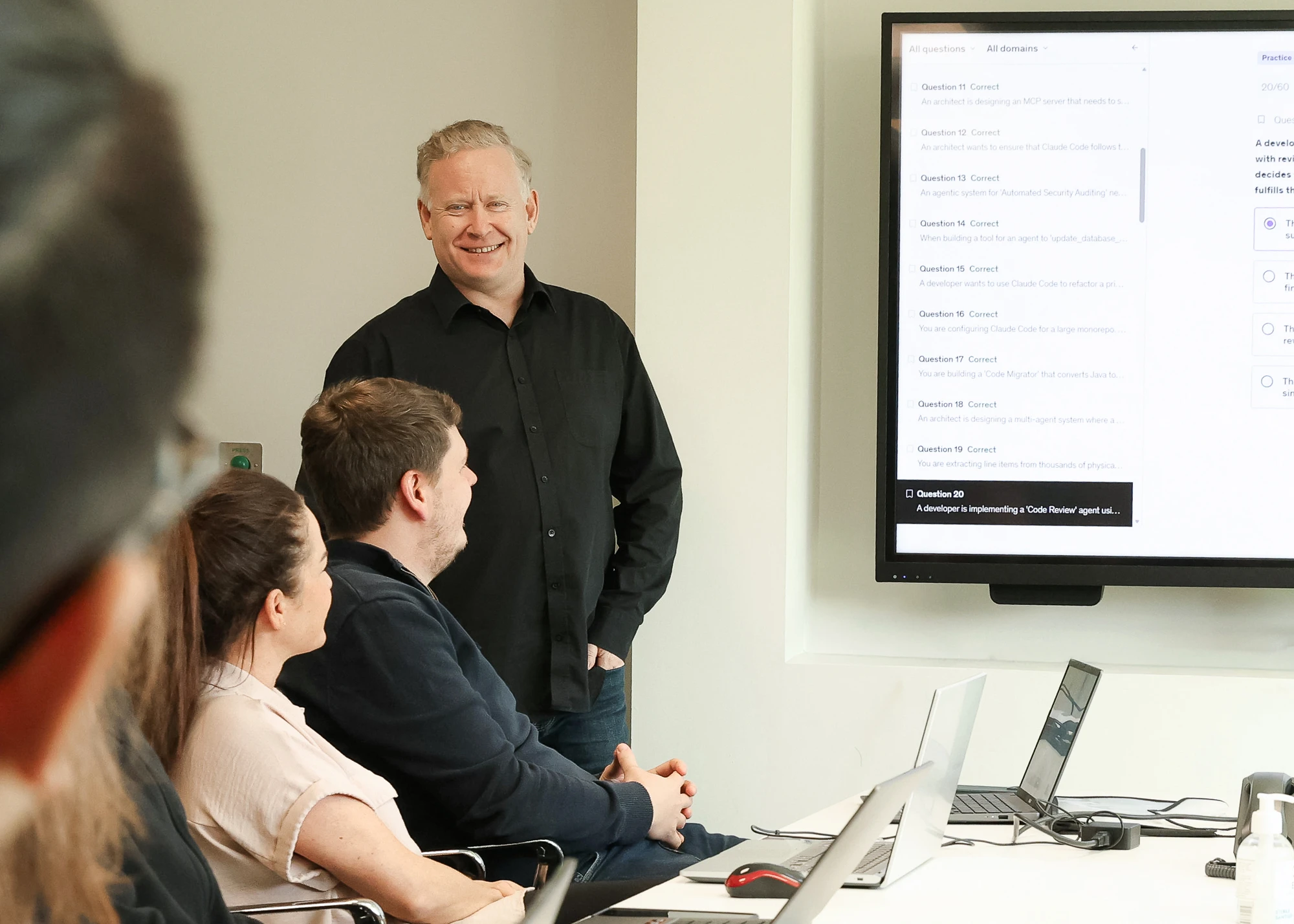 A man in a black shirt smiles while presenting information on a large screen to three seated people in a meeting room with laptops on the table.