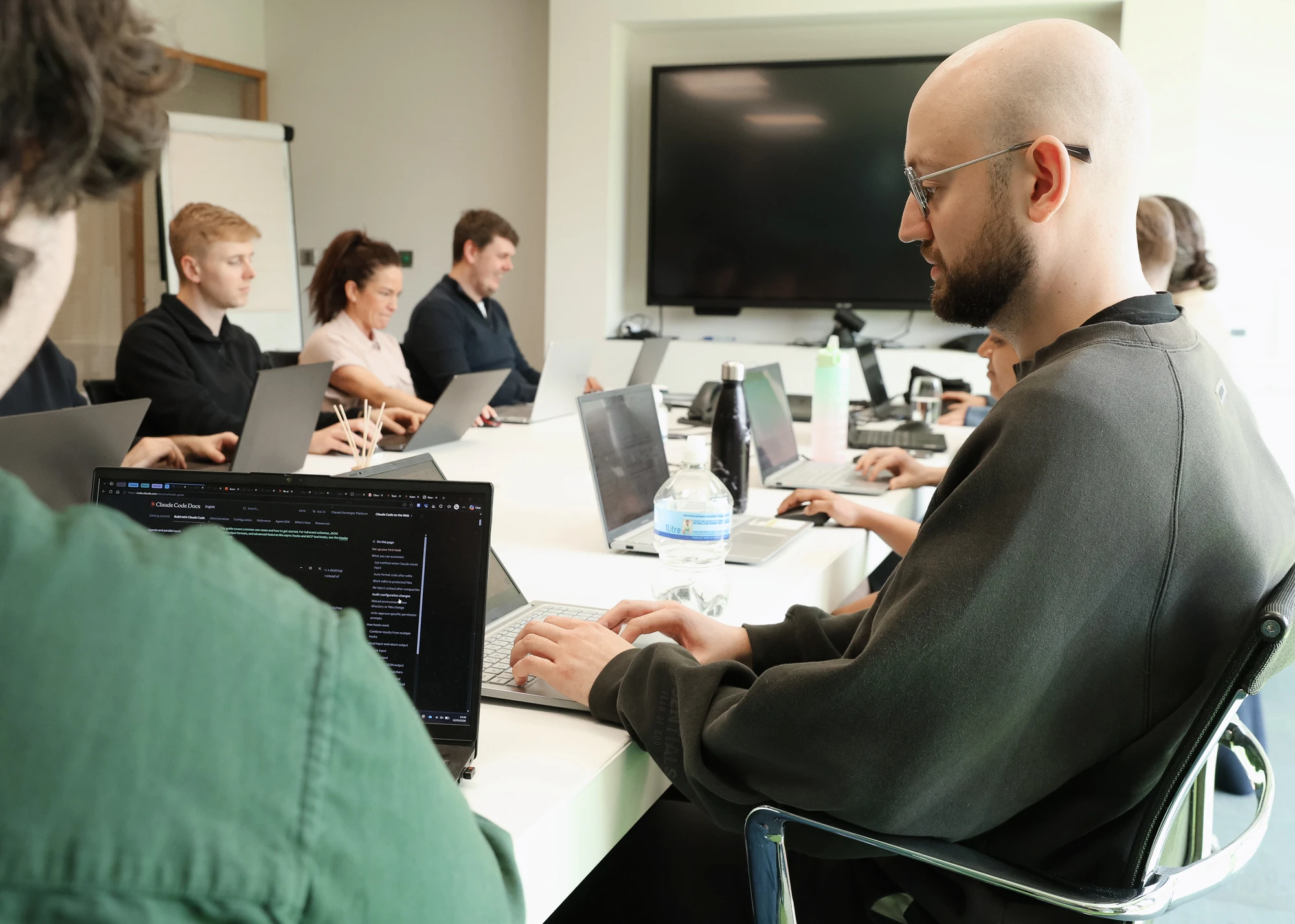 A group of people sitting around a white conference table in a meeting room, each working on laptops, with a large TV screen on the wall in the background and water bottles on the table.