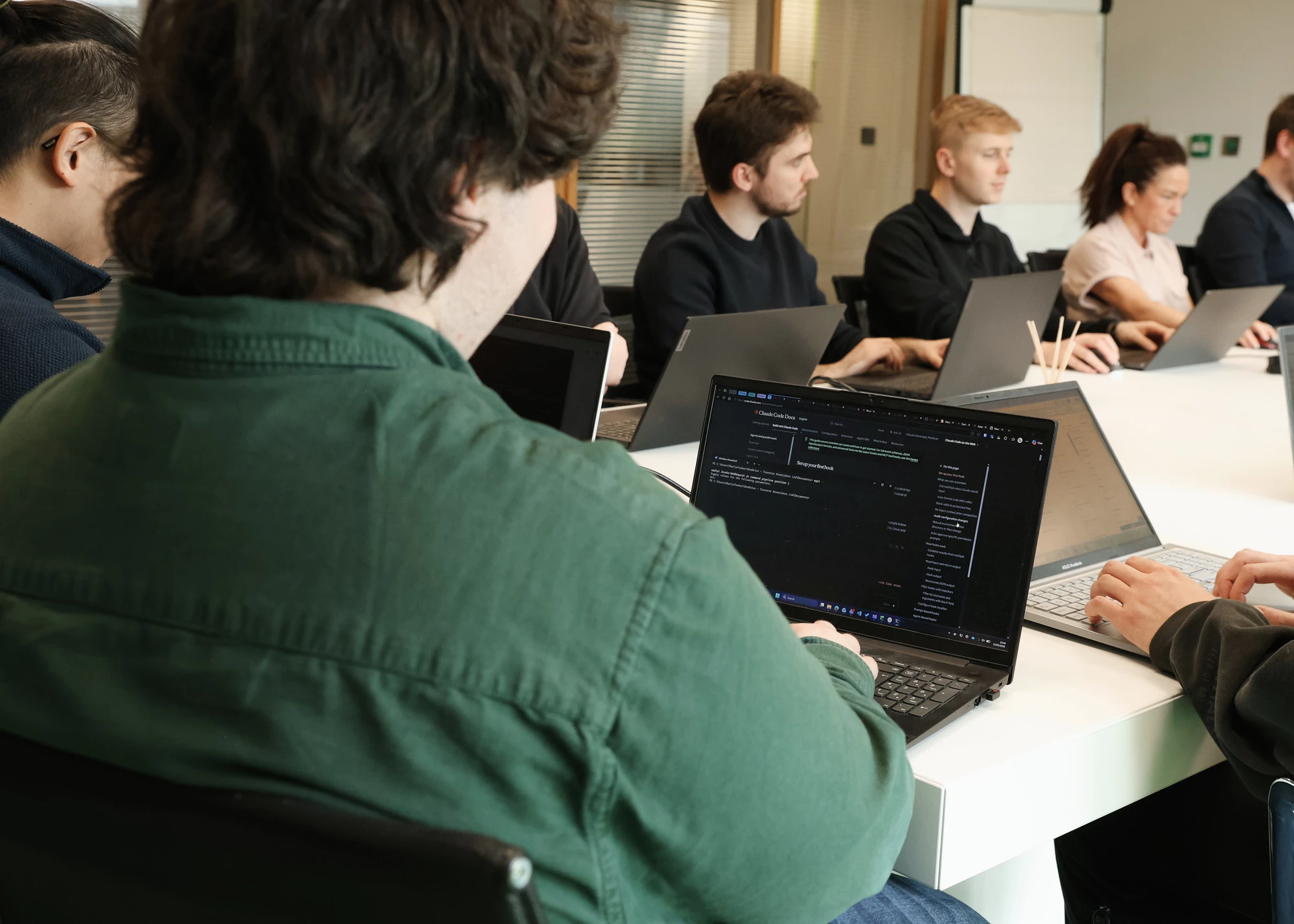 Group of people sitting around a white table working on laptops, focused on screens showing code and documents in an office setting.