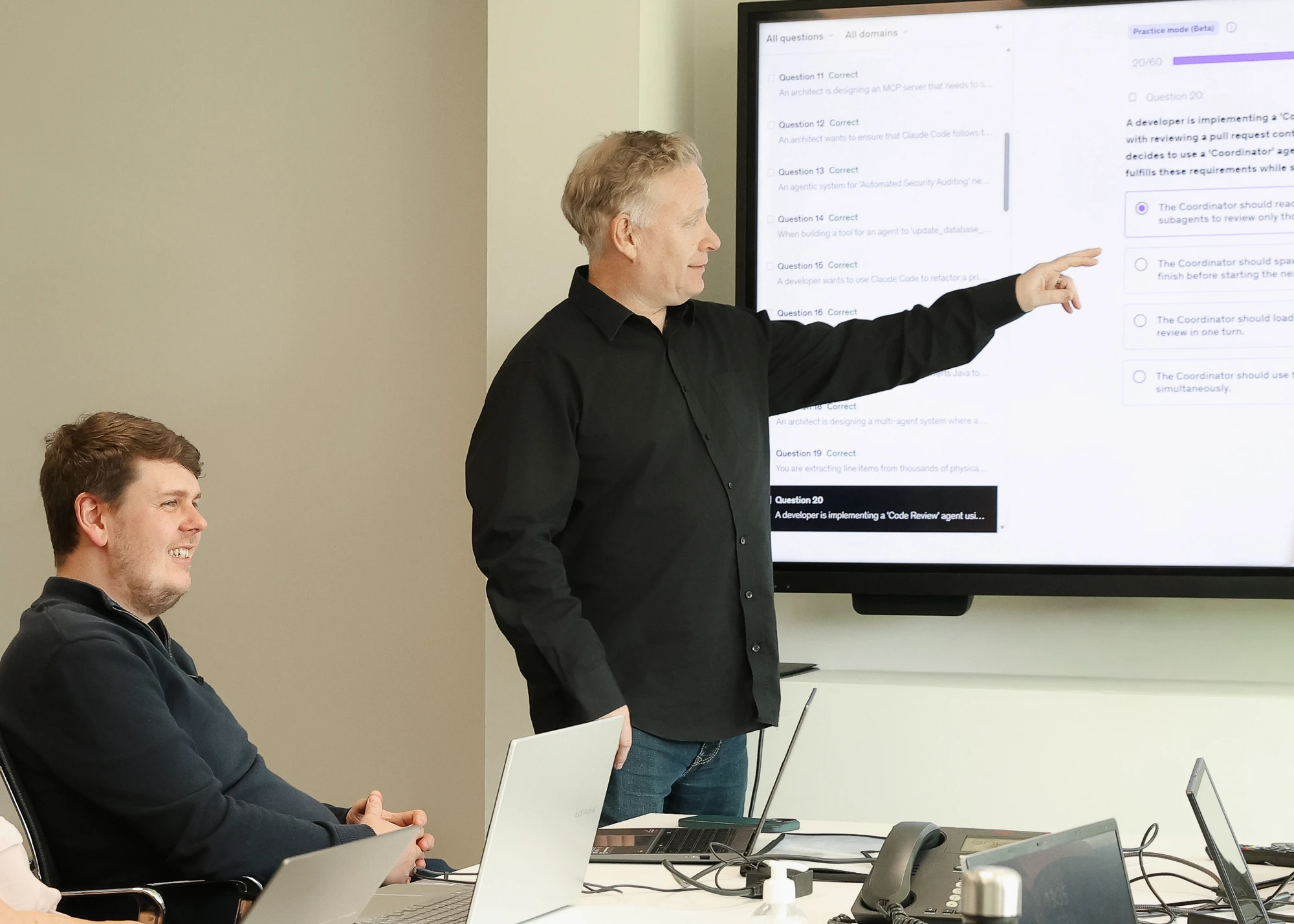 A man in a black shirt standing and pointing at a large screen displaying a multiple-choice quiz interface, while another man sitting at the table with laptops listens and smiles.
