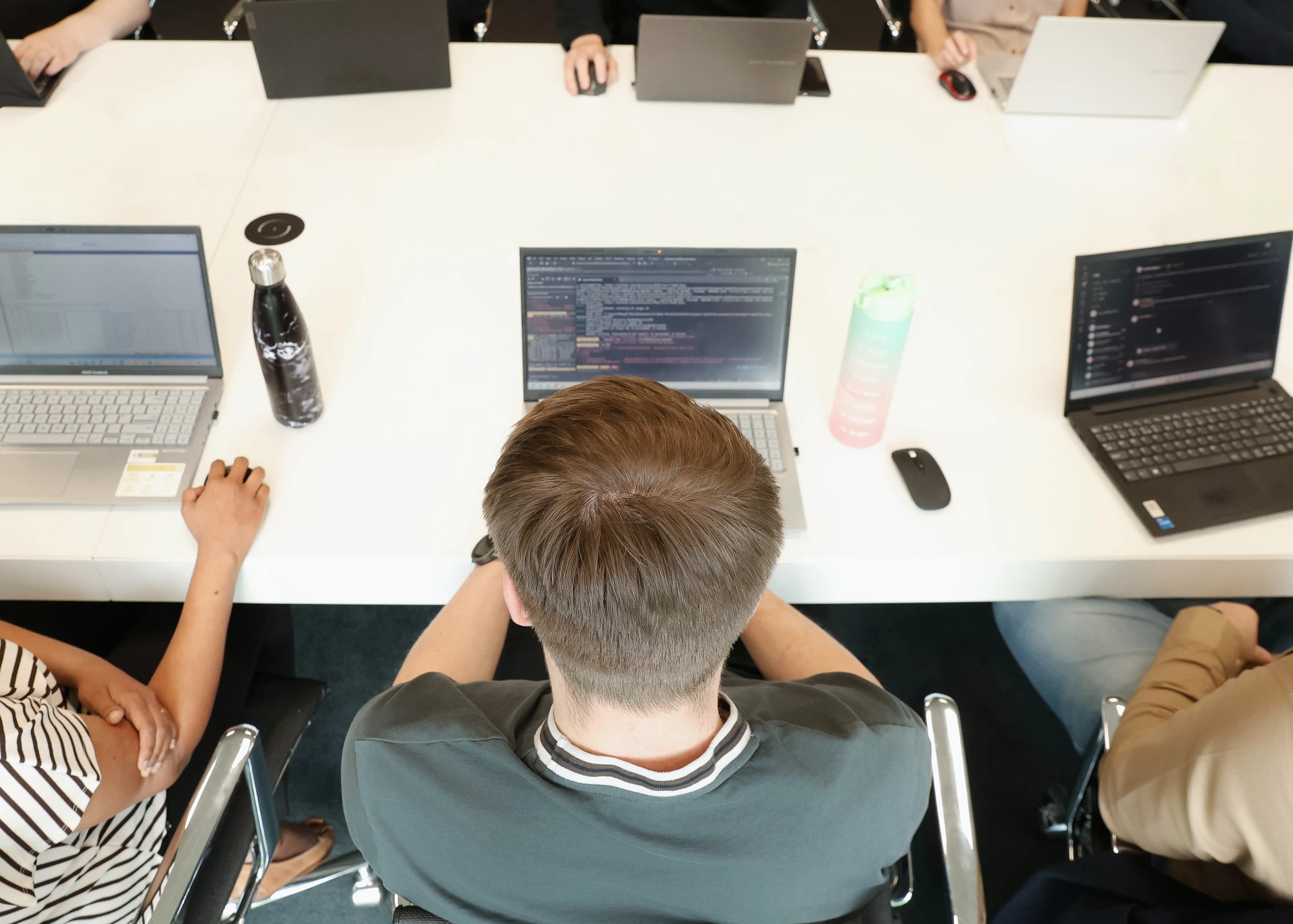 Overhead view of six people sitting at a white table working on laptops with water bottles and computer mice scattered on the table.