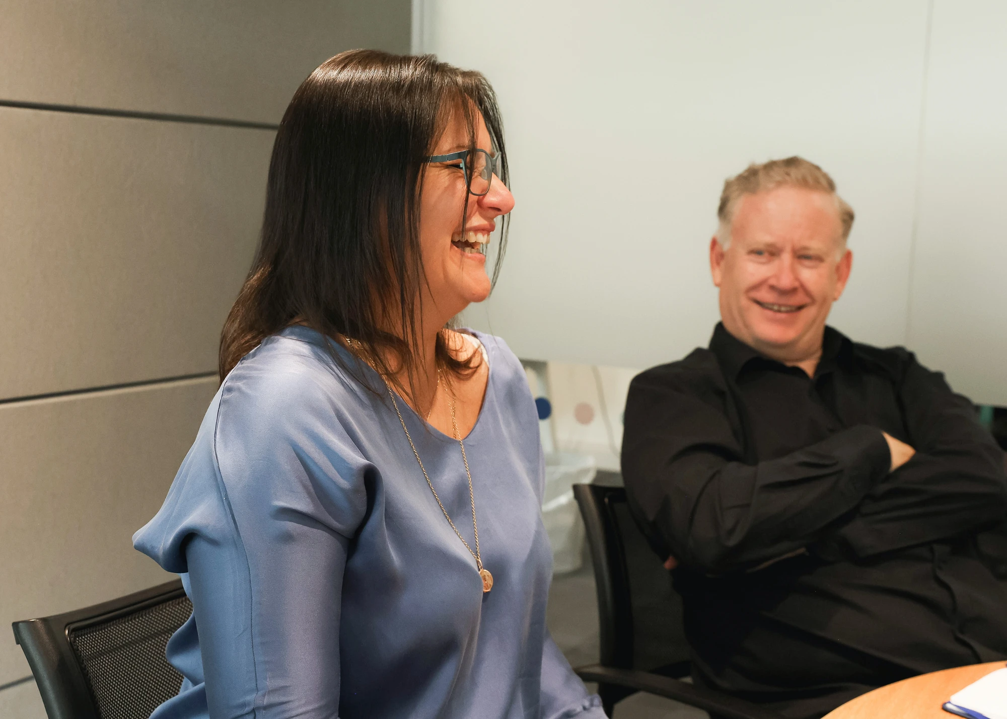 Two people seated in chairs in an office setting; a woman with long brown hair and glasses wearing a blue blouse is laughing, and a man with short blonde hair wearing a black shirt is smiling with his arms crossed.
