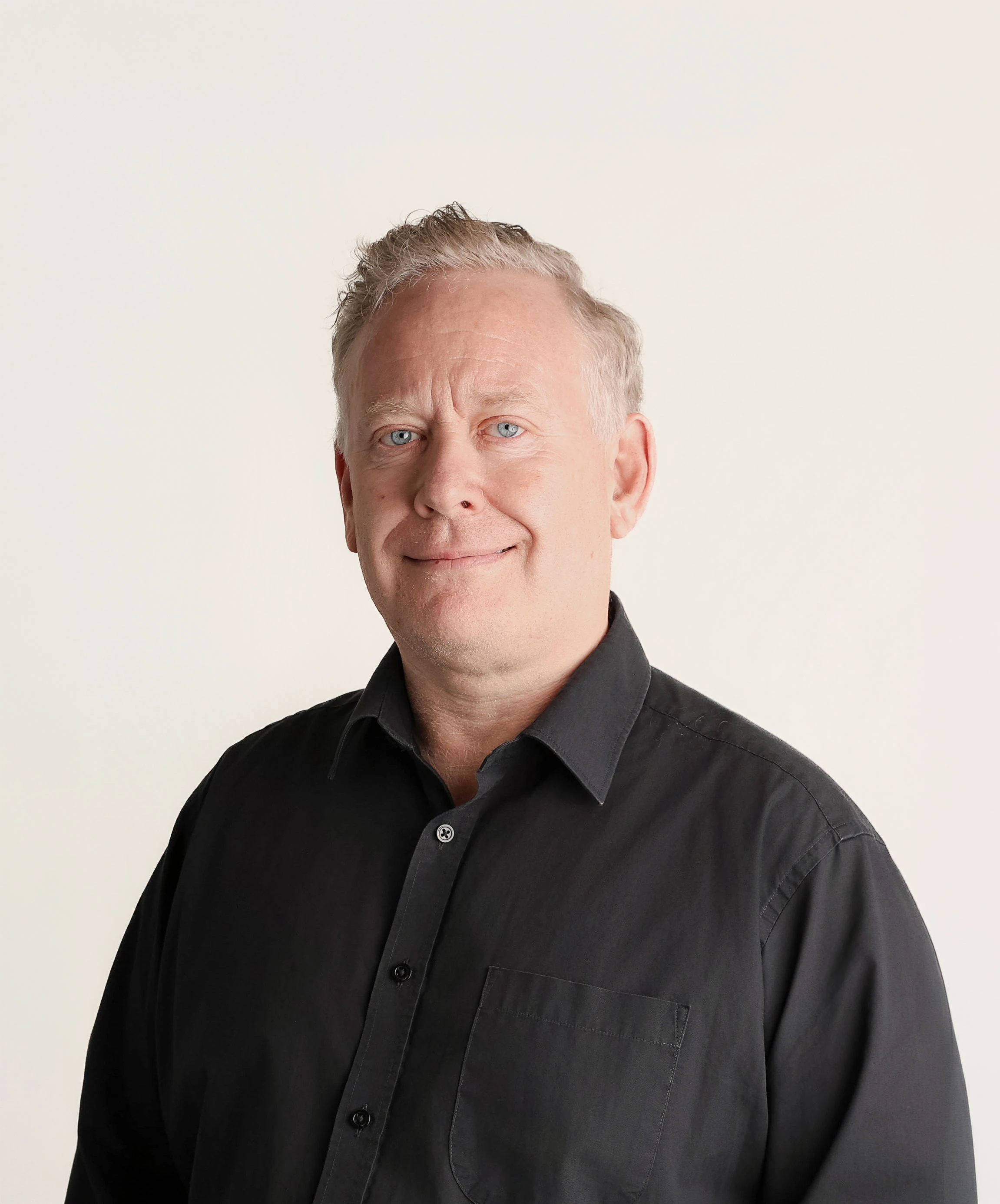 Portrait of a man with light skin, short light hair, and blue eyes, wearing a black button-up shirt, smiling slightly against a plain light background.