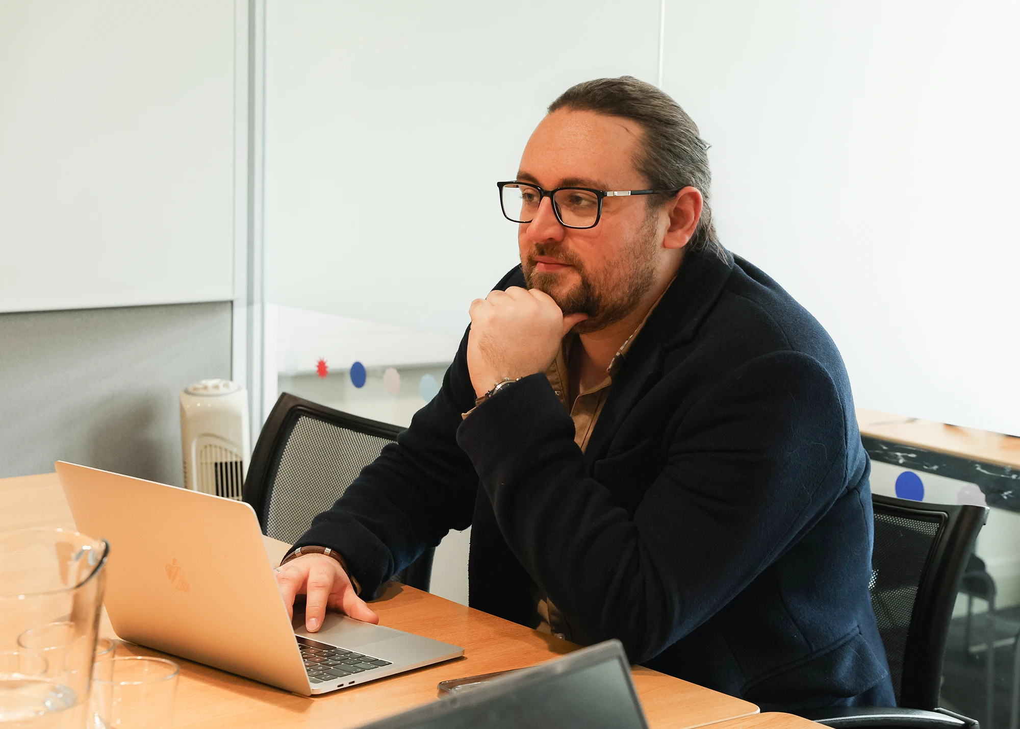 Bearded man with glasses and long hair tied back, wearing a dark jacket, sitting at a wooden table working on a silver MacBook laptop in a modern office setting.