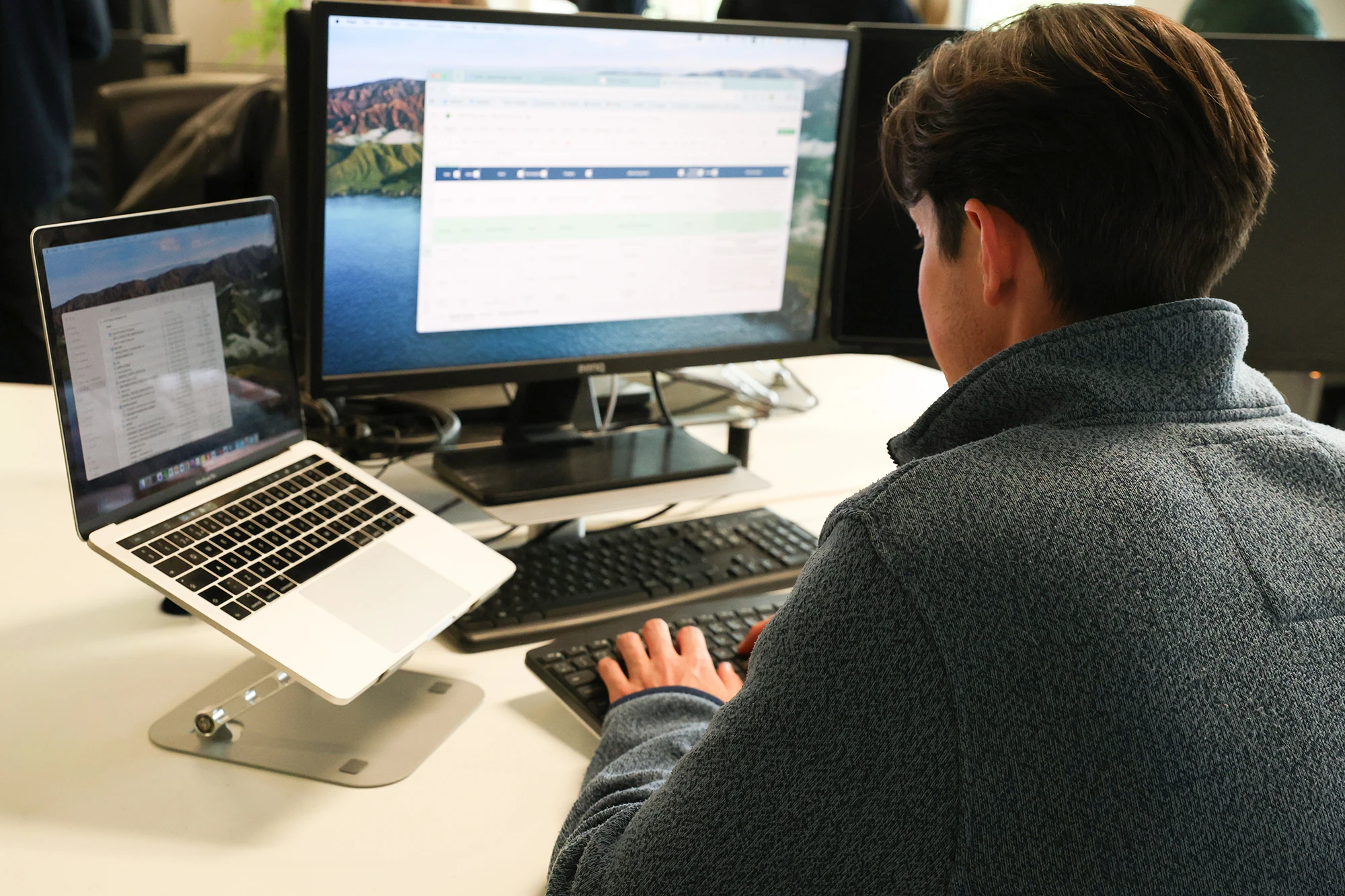 Person in a gray sweater working on a desktop computer with a monitor and a laptop on a stand, typing on a keyboard at a white desk.