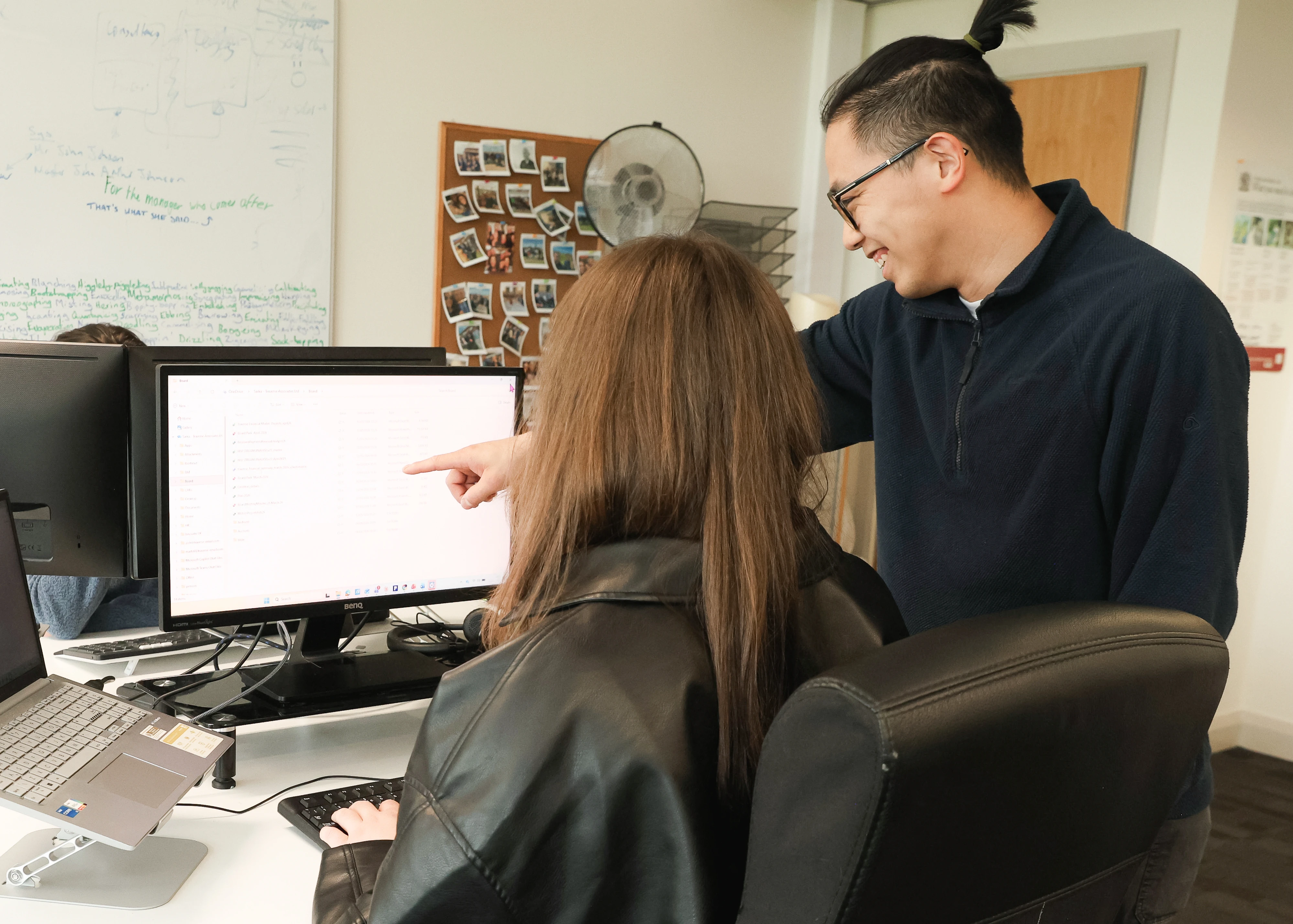 A man with glasses and a navy sweater stands and smiles while pointing at a computer screen, and a woman with long brown hair in a black leather jacket types on a keyboard in an office setting.
