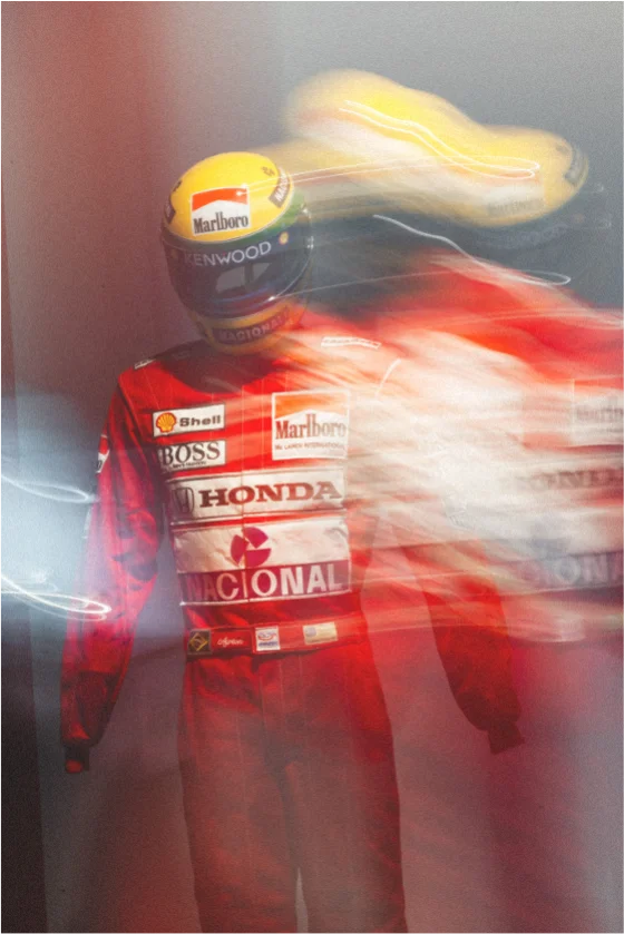Blurred image of a racing driver in a red racing suit with logos including Marlboro, Honda, Shell, and Boss, wearing a yellow helmet with Marlboro branding, creating a motion effect.