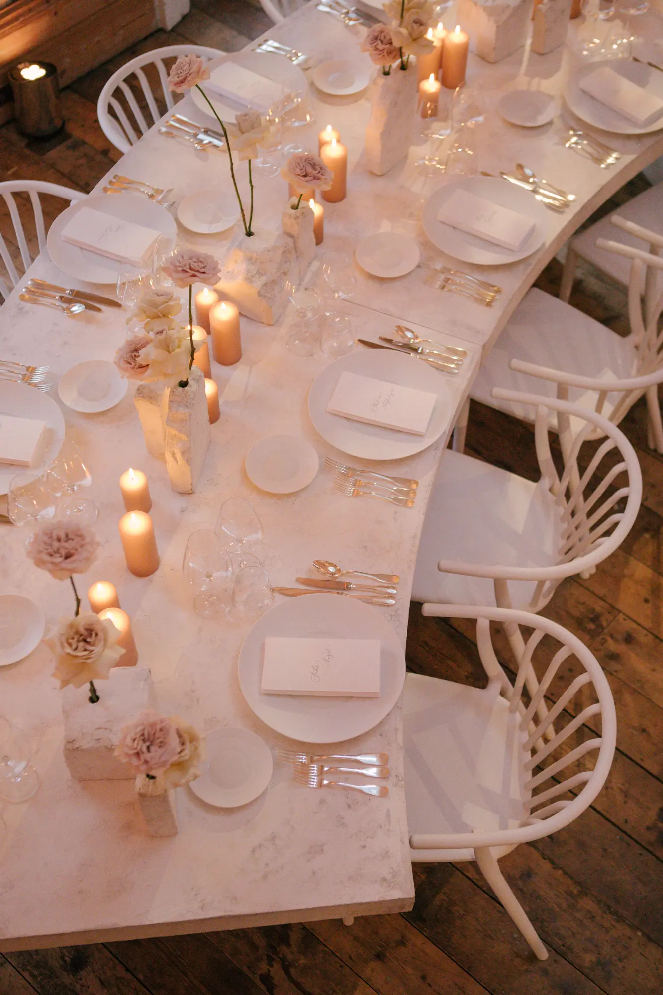 Elegant dining table set with white plates, menus, silver cutlery, wine glasses, and soft pink flowers in rustic stone vases, illuminated by lit candles, surrounded by white chairs on a wooden floor.