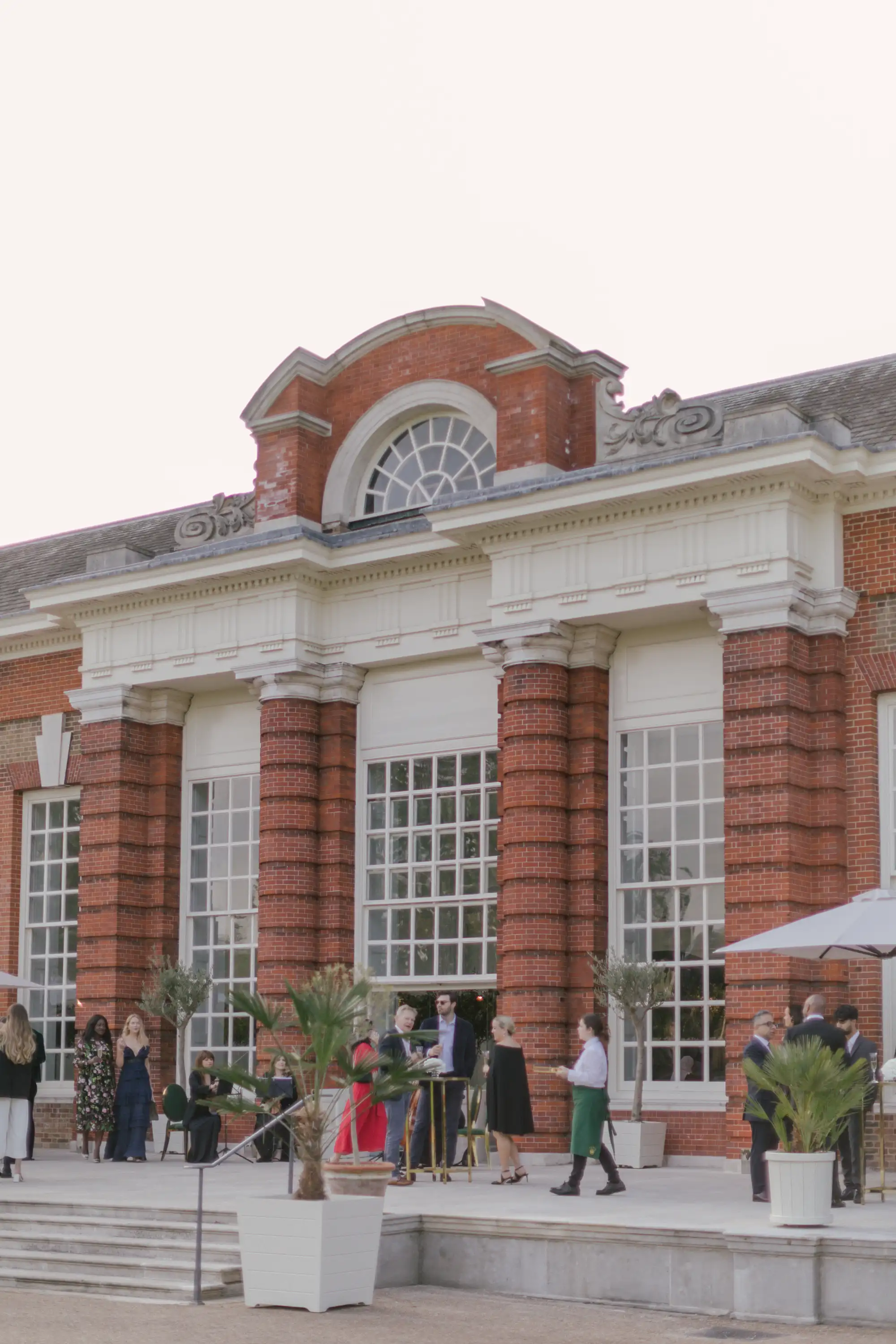 Elegant outdoor social gathering with people dressed in formal attire standing and sitting near a large brick building with tall windows and decorative architectural details.