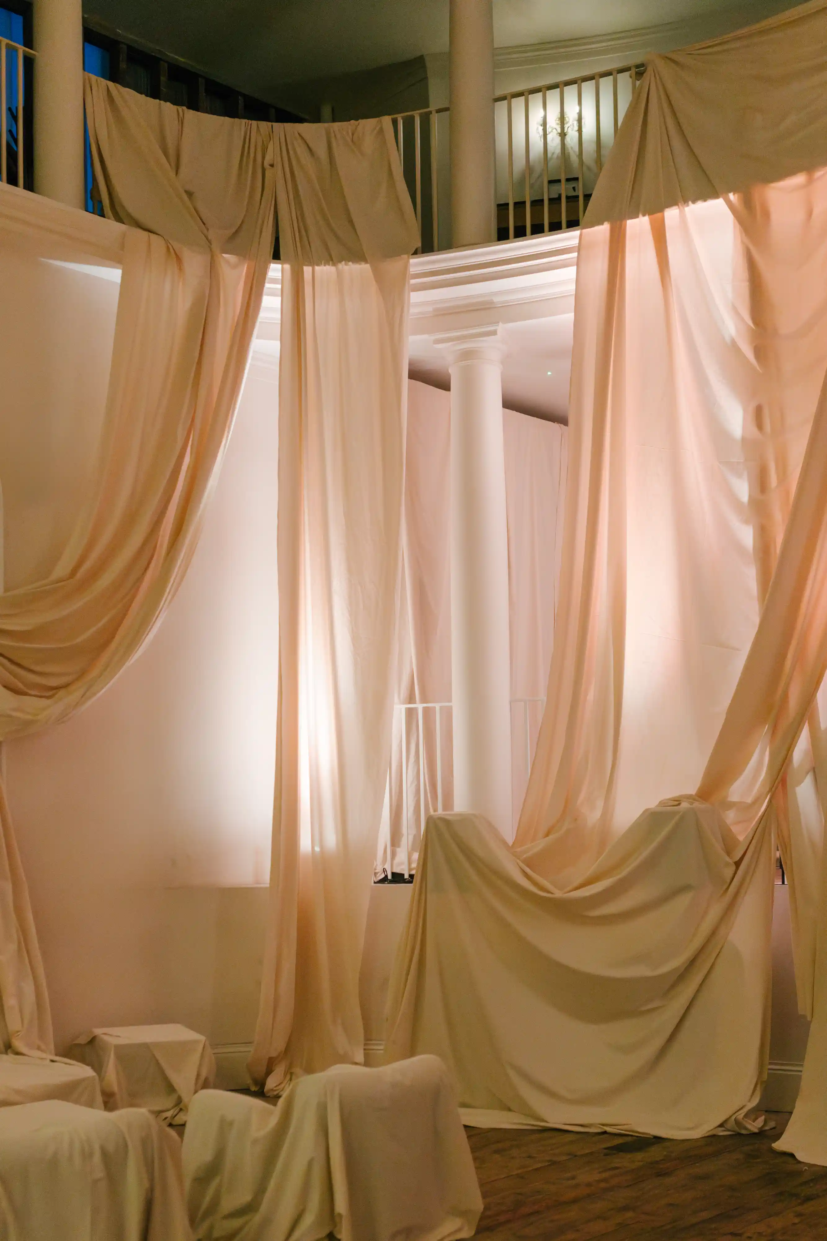 Interior space with tall white columns and beige fabric draped from the ceiling and covering furniture, softly lit with warm light.
