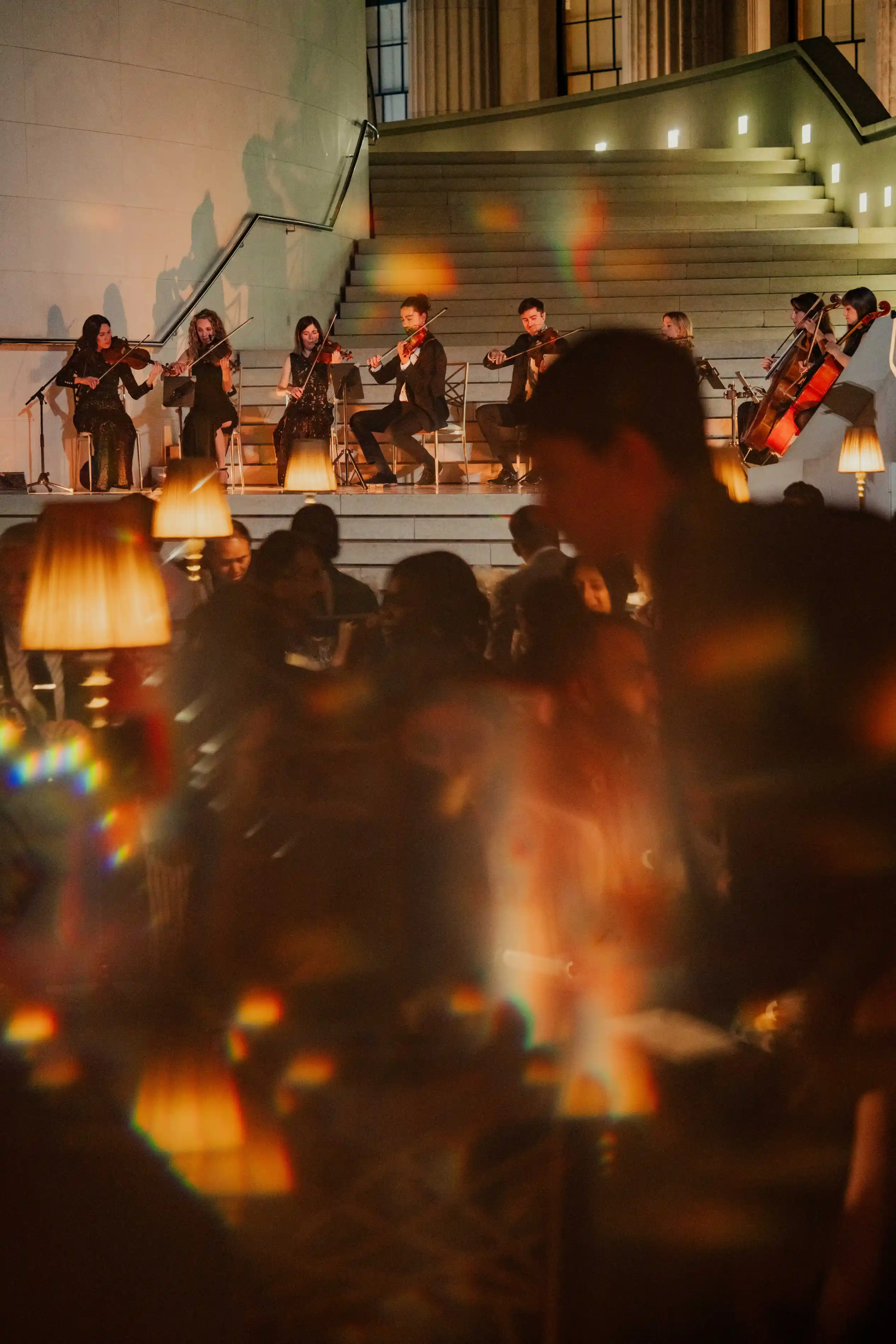Evening indoor scene with a string ensemble of seven musicians playing violins and cellos seated on steps, illuminated by soft lights, while blurred silhouettes of people and warm amber lamps fill the foreground.