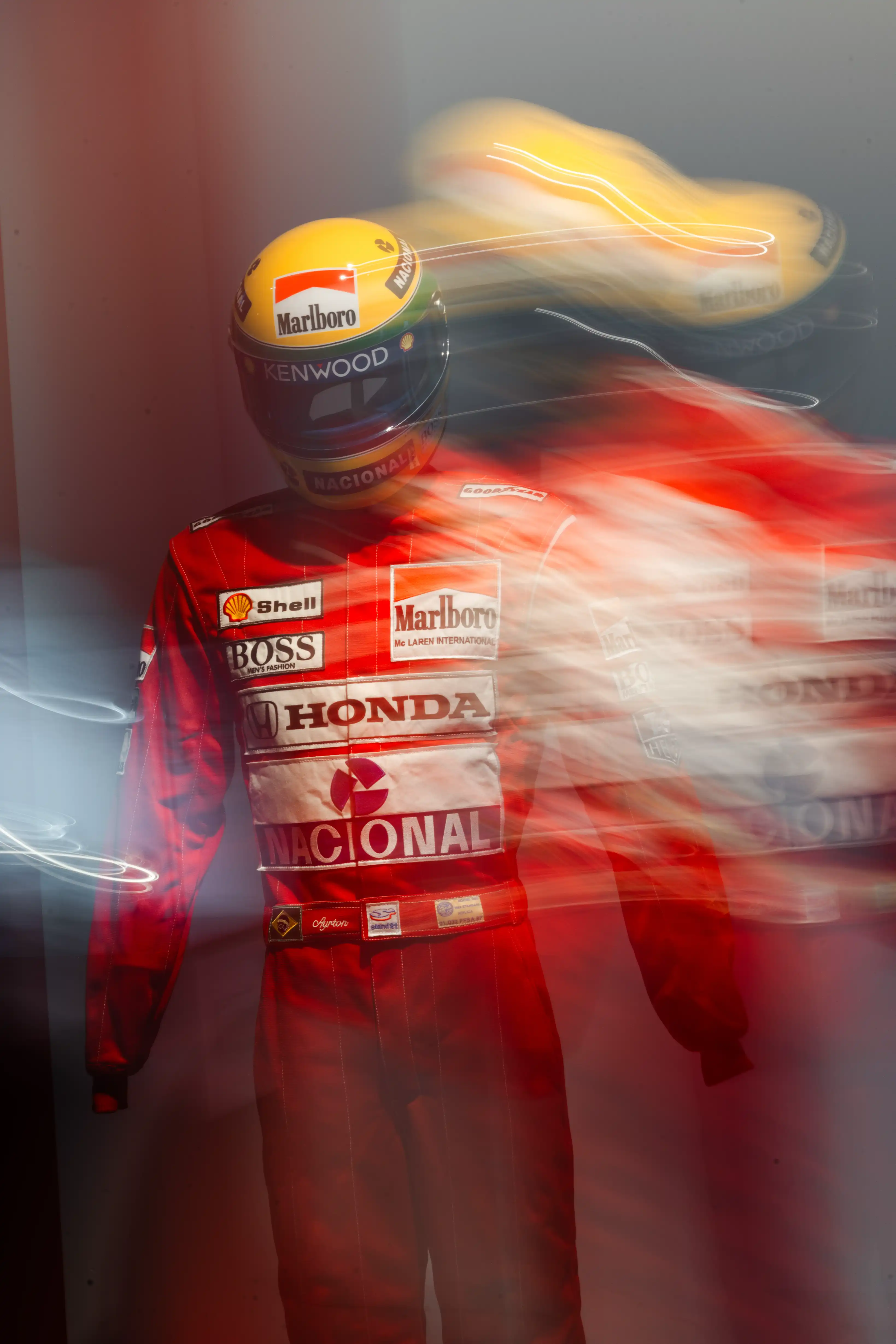 Figure in a red racing suit with sponsor logos including Marlboro, Shell, Boss, Honda, and Nacional, wearing a distinctive yellow helmet, captured with a motion blur effect creating ghost-like multiple images to the right.