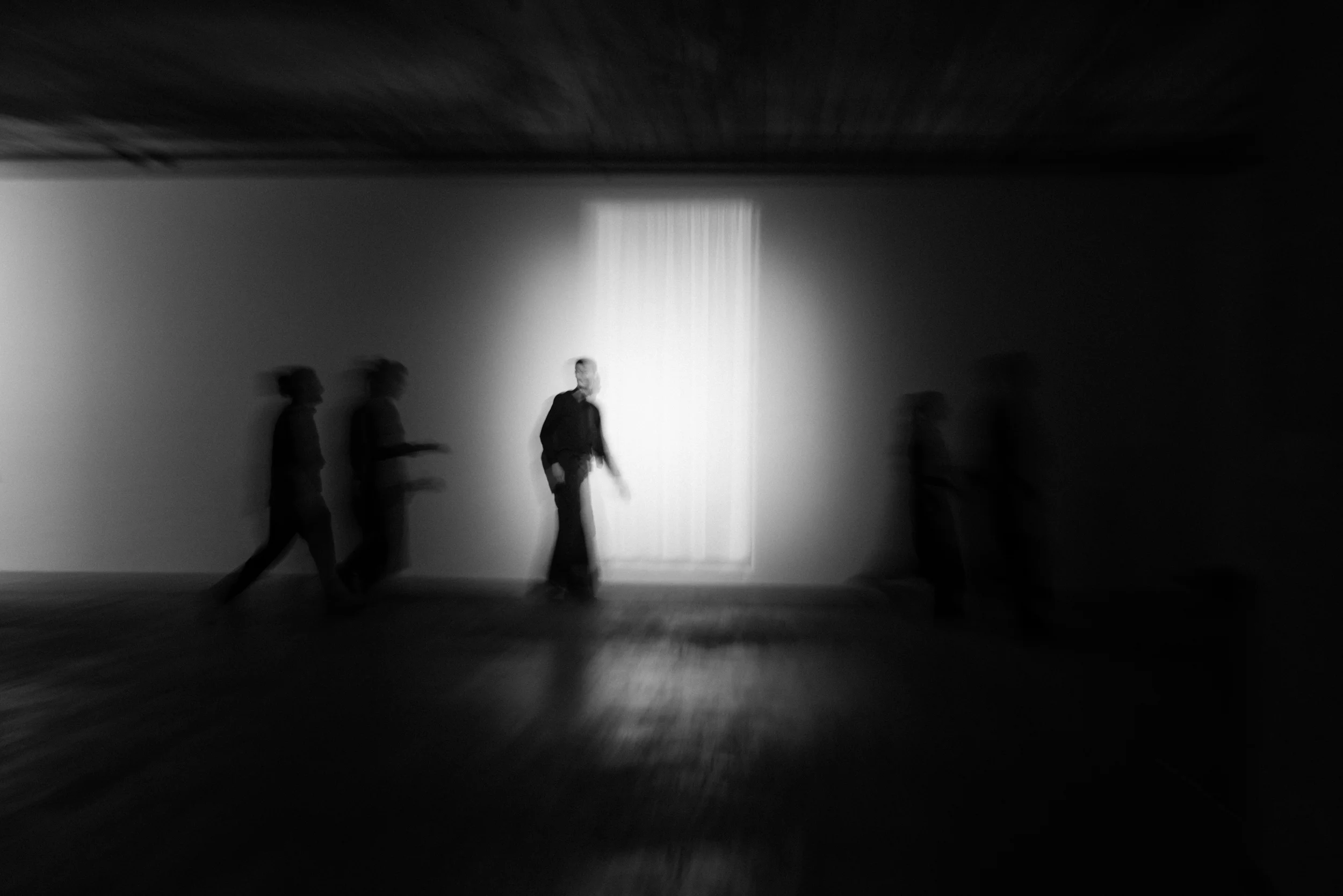 Blurry black-and-white photo showing multiple ghostly silhouettes of a person walking past a bright window on a dark wall.