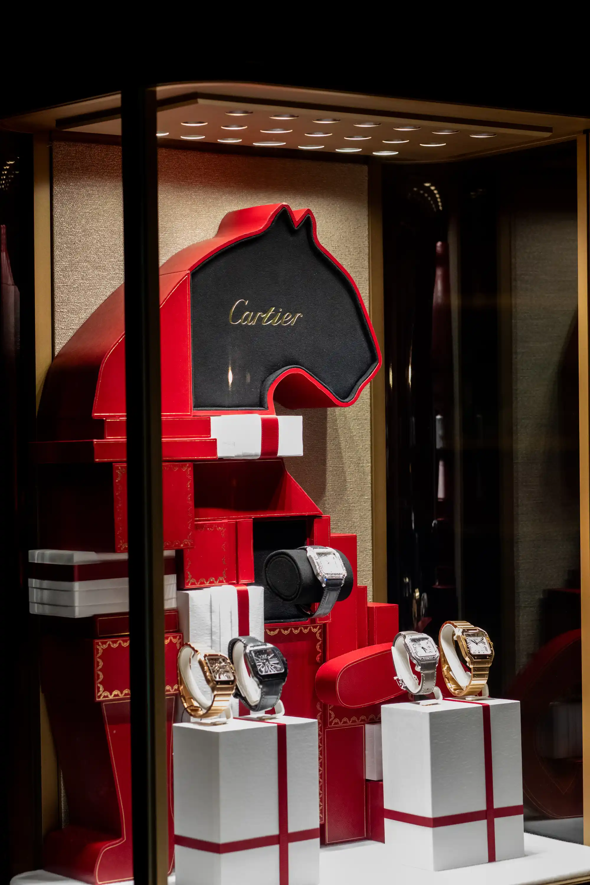 Cartier watch display in a store window featuring five luxury wristwatches on white podiums with red ribbon accents, set against a backdrop shaped like a horse's head in red and black with the Cartier logo in gold.