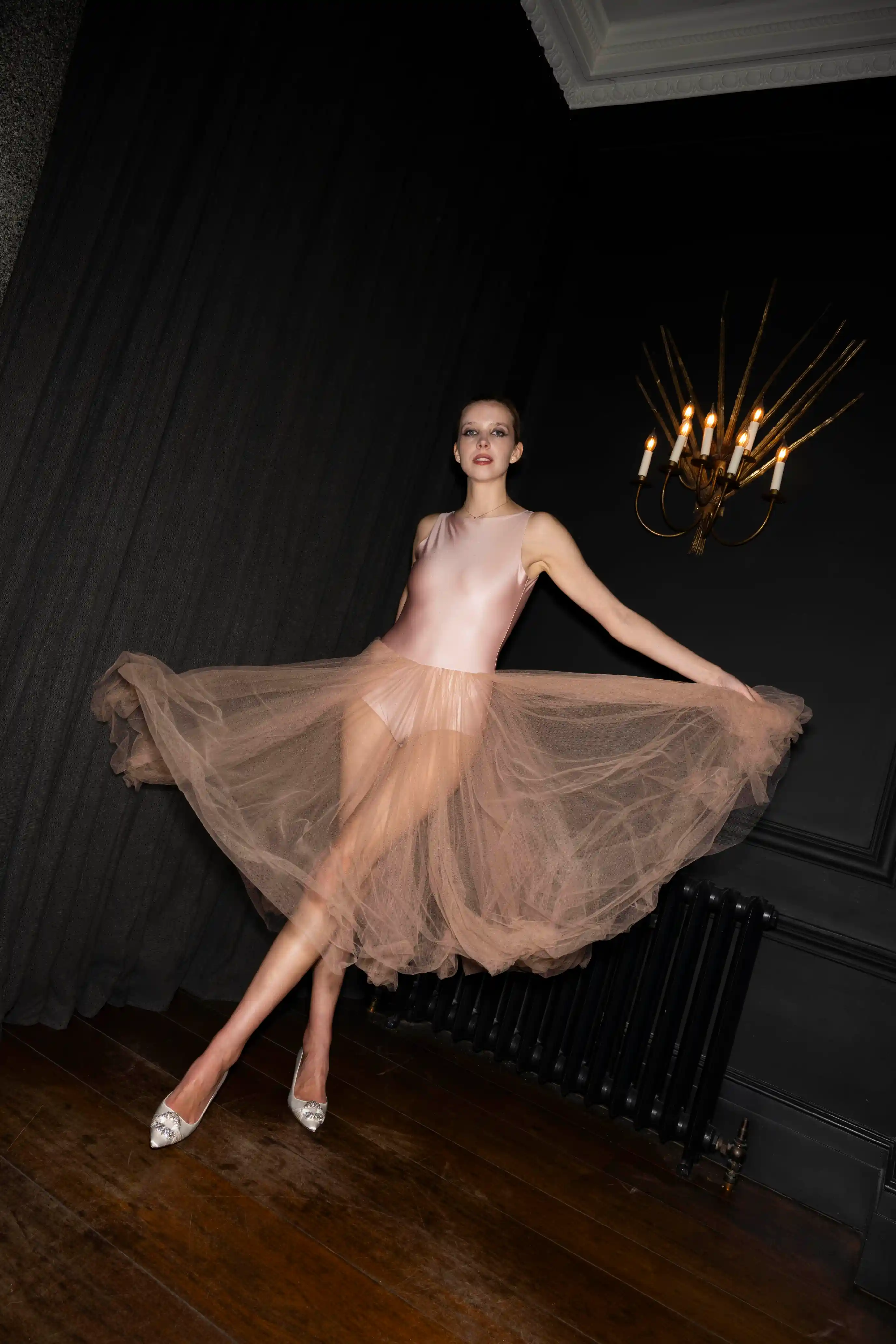A woman wearing a pink satin leotard and a sheer flowing tulle skirt poses gracefully on a wooden floor in a dark room with black walls and a vintage chandelier with lit candles.