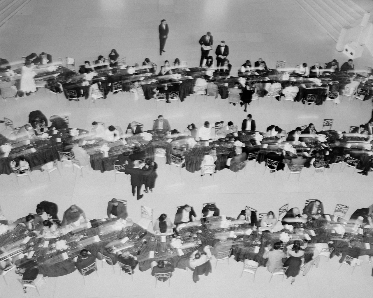 Black and white overhead photo of a formal event with three long, curved banquet tables crowded with seated guests dressed in formal attire, some interacting and a few standing on the large open floor space.