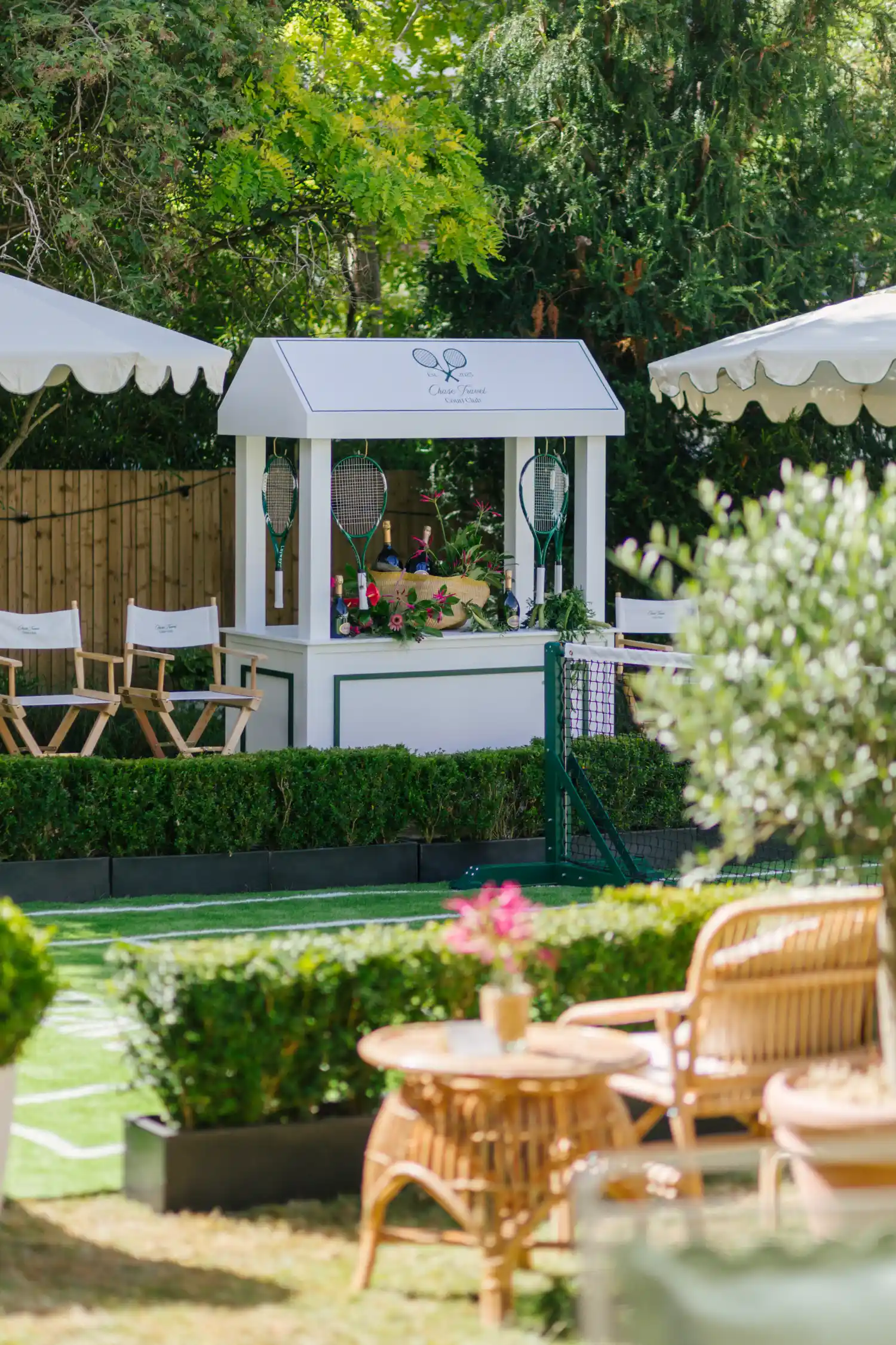 Outdoor tennis court scene featuring a white refreshments stand decorated with tennis rackets and floral arrangements, surrounded by director chairs under white canopies, green hedges, and wicker furniture with a small table and pink flowers in the foreground.