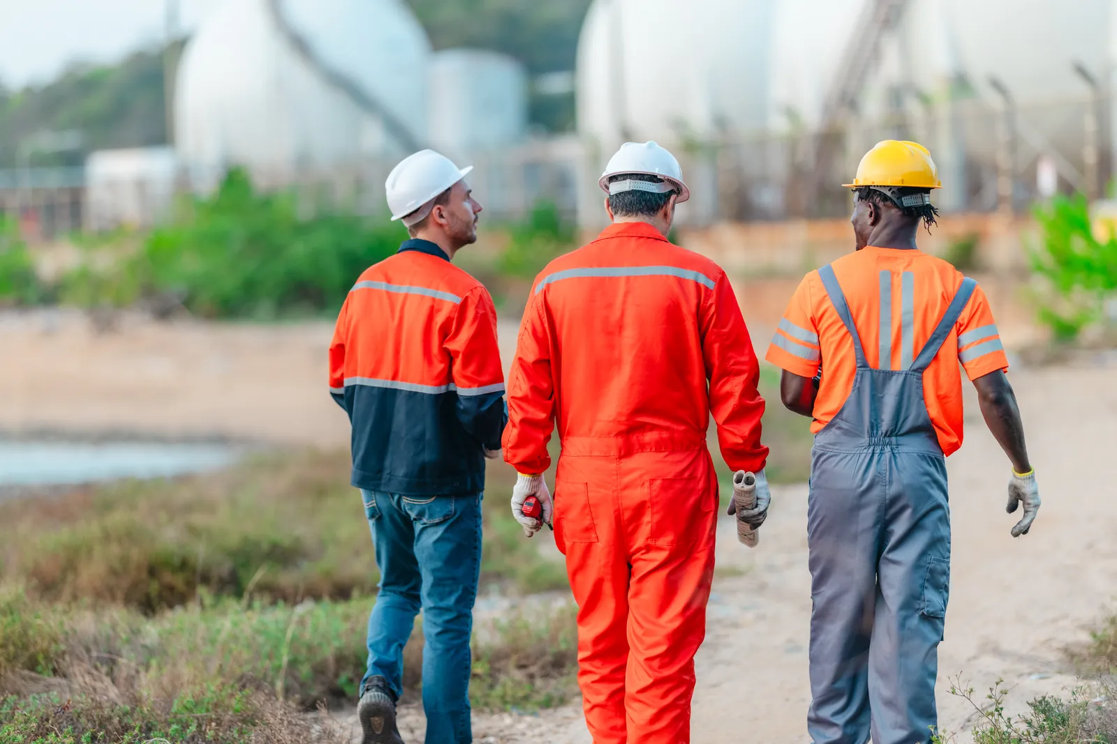Three workers in coveralls and hard hats walking toward storage tanks
