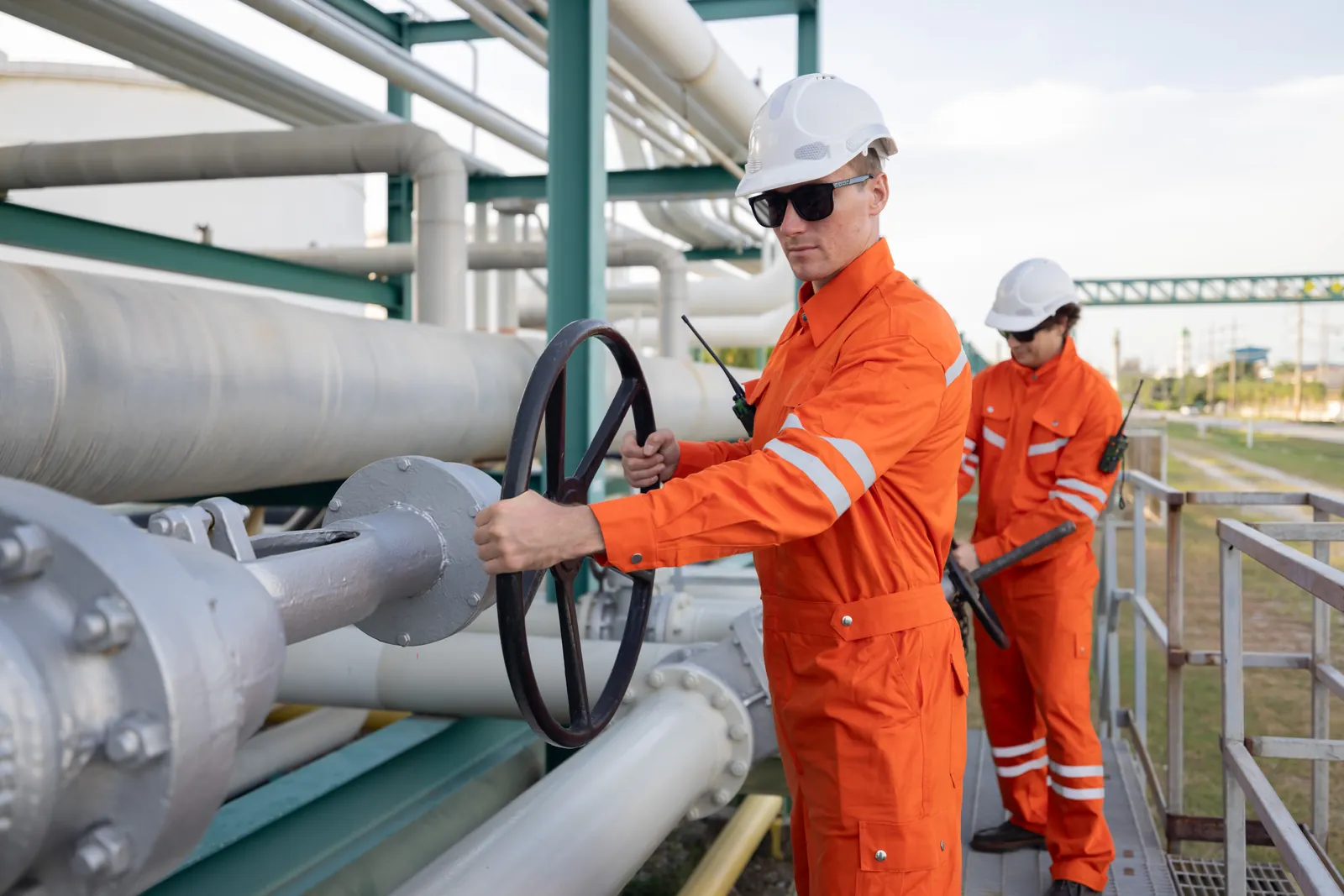 Workers in orange coveralls turning a valve on large piping