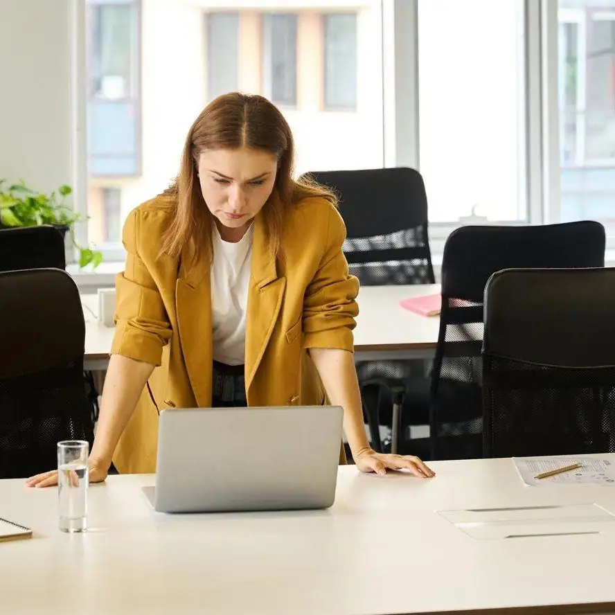 A team member in a mustard blazer leaning over a laptop at a white desk in a bright office