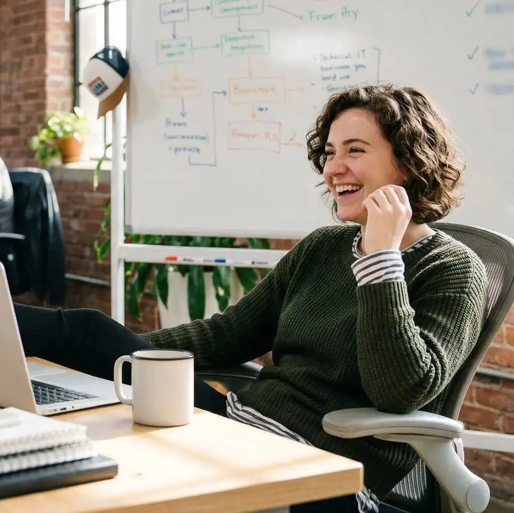 A smiling team member with curly hair, in an olive knit sweater, laughing at her desk in front of a whiteboard