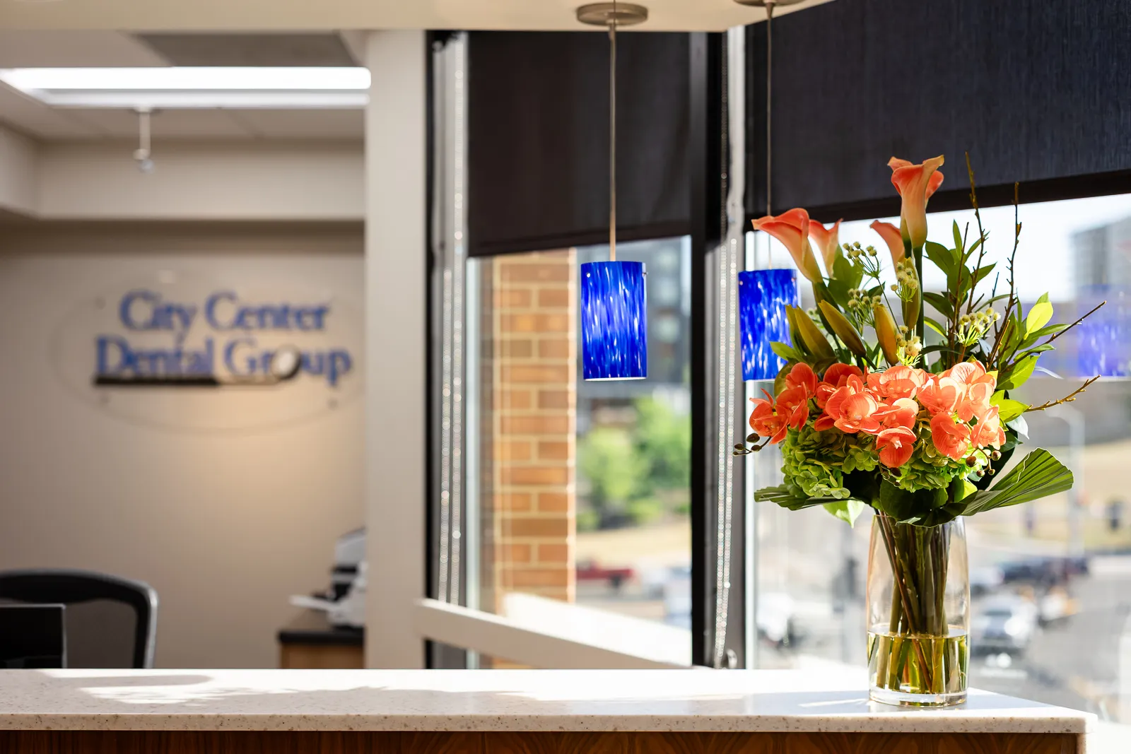Reception desk with a clear glass vase of orange and green flowers, in front of a window with black blinds and blue hanging pendant lights, with a blurred City Center Dental Group sign in the background.