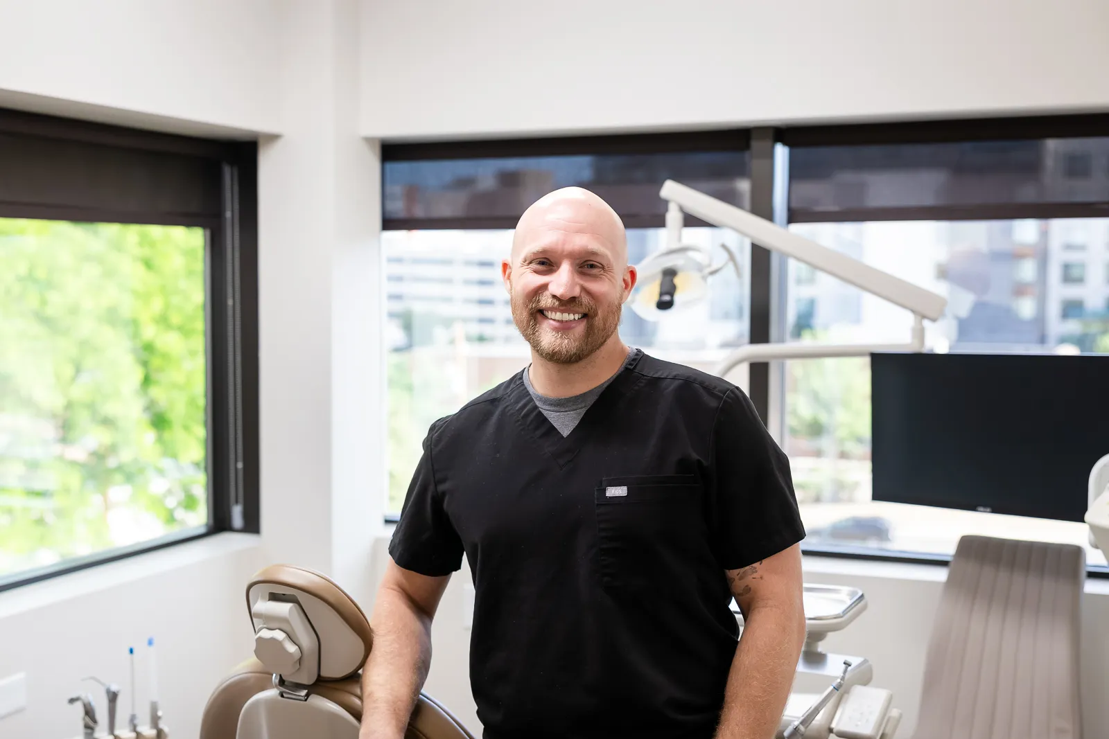Smiling bald man with a beard wearing black scrubs standing in a bright dental office with dental chair and equipment near large windows showing city buildings outside.