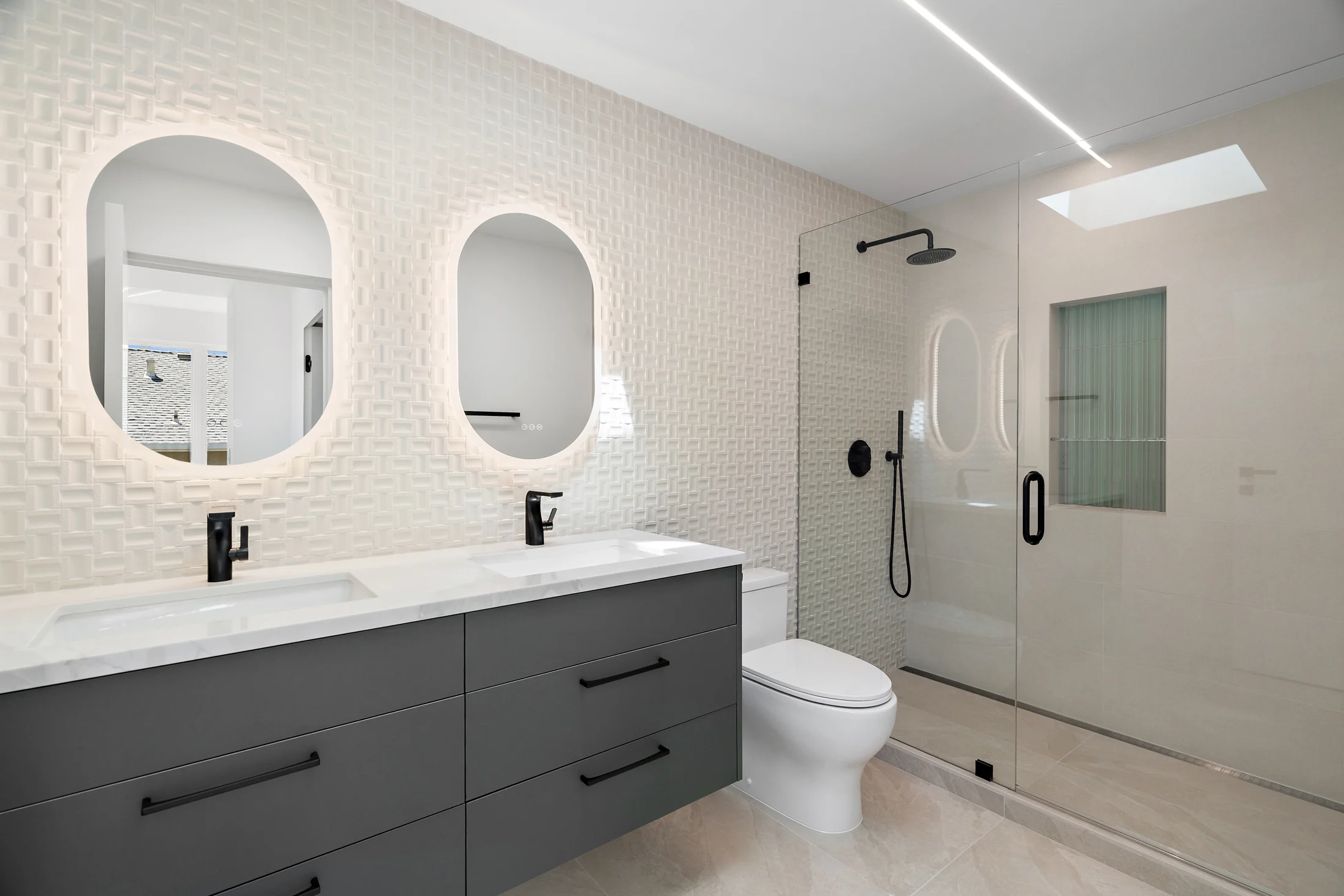 Bathroom with a double vanity, oval mirrors, glass shower and skylight.