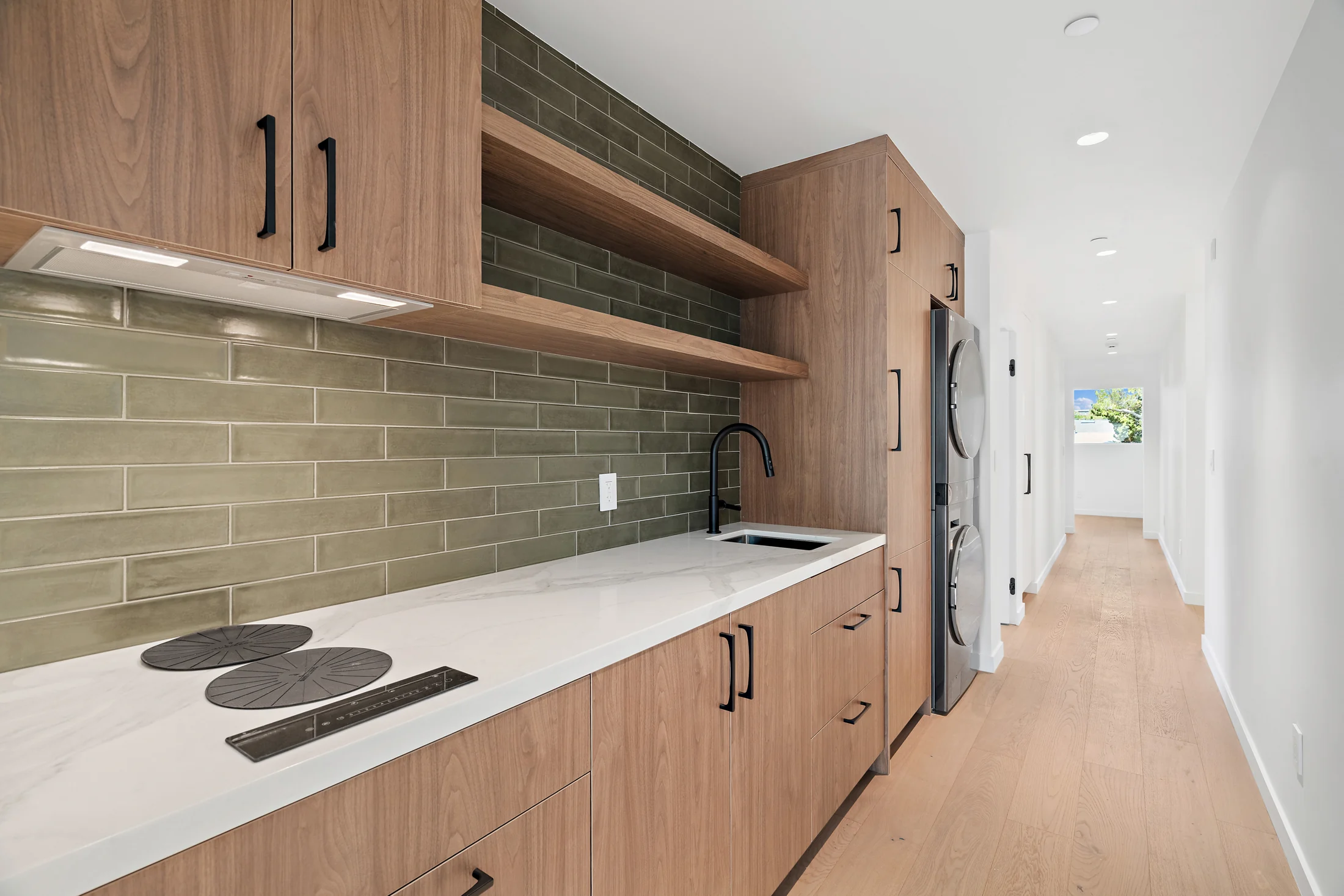 ADU kitchenette with wood cabinetry, green tile backsplash and stacked laundry appliances.