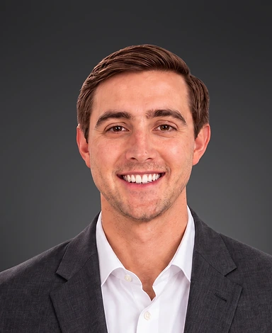 Smiling young man with short brown hair wearing a dark gray suit jacket and a white dress shirt against a dark gray background.
