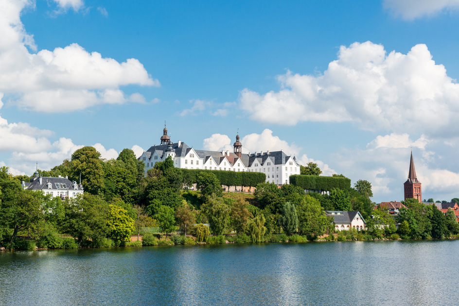 Das Plöner Schloss liegt eingebettet in der wunderschönen Seenlandschaft der Holsteinischen Schweiz. Der Plöner See im Vordergrund bei Sonnenschein.